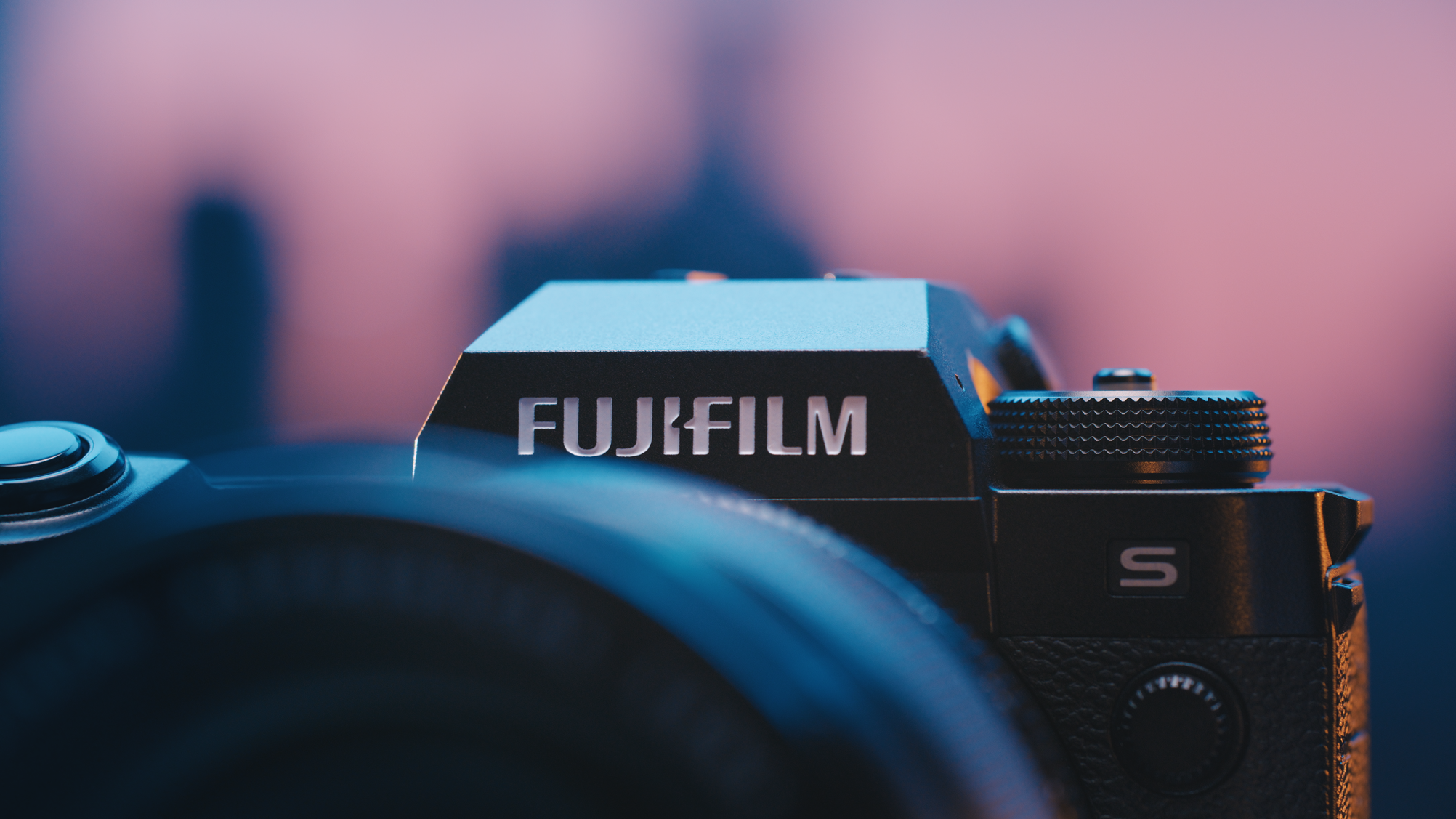 Close-up of a Fujifilm camera with textured dials, against a pink and purple gradient background.