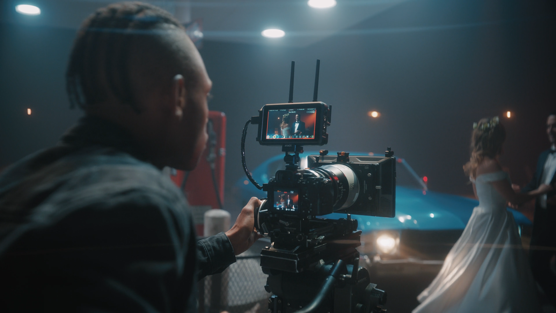 A film crew filming a scene with a man and woman in formal attire in front of a blue sports car on a dimly lit street.