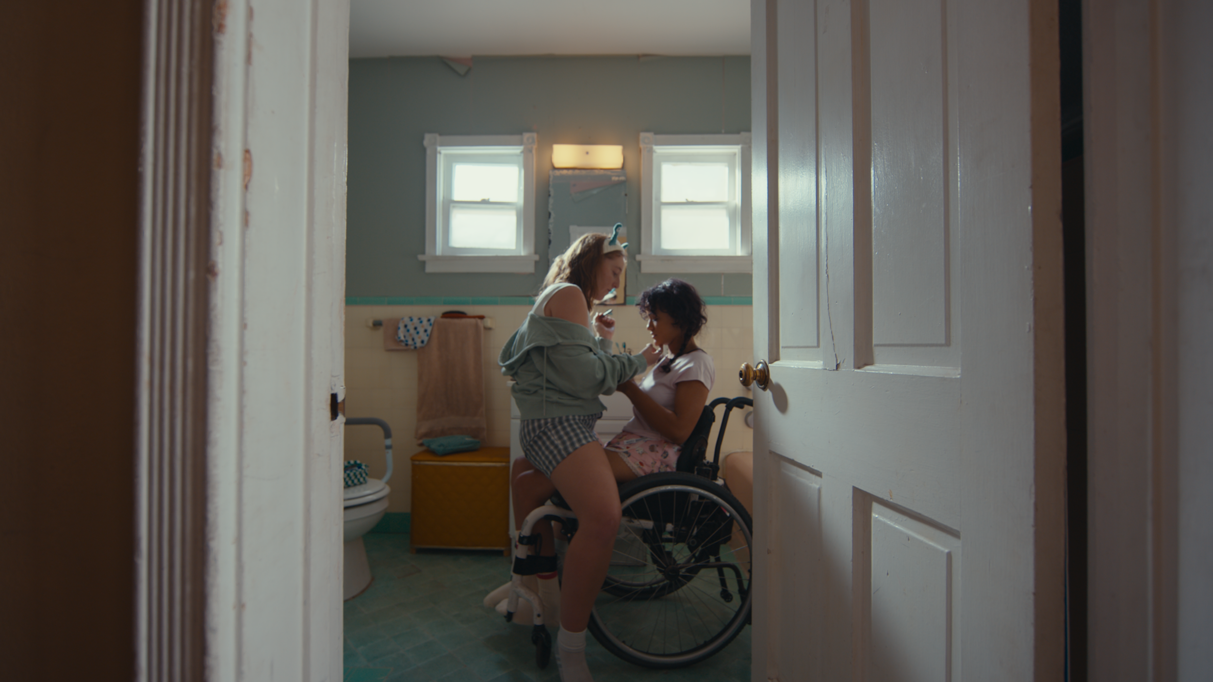 A woman with a headband assisting a girl in a wheelchair in a bathroom.