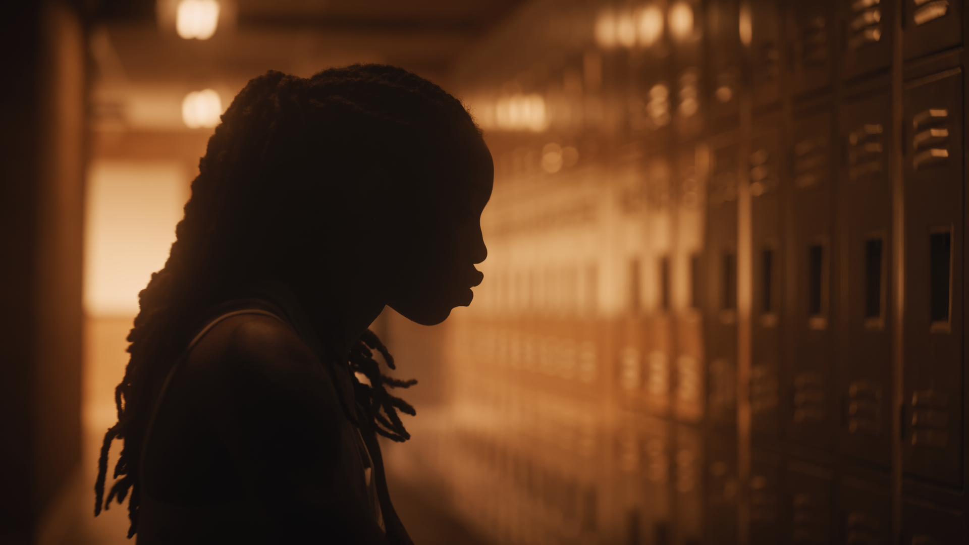 Silhouette of a young woman with dreadlocks standing in front of a row of lockers in a dimly lit hallway.