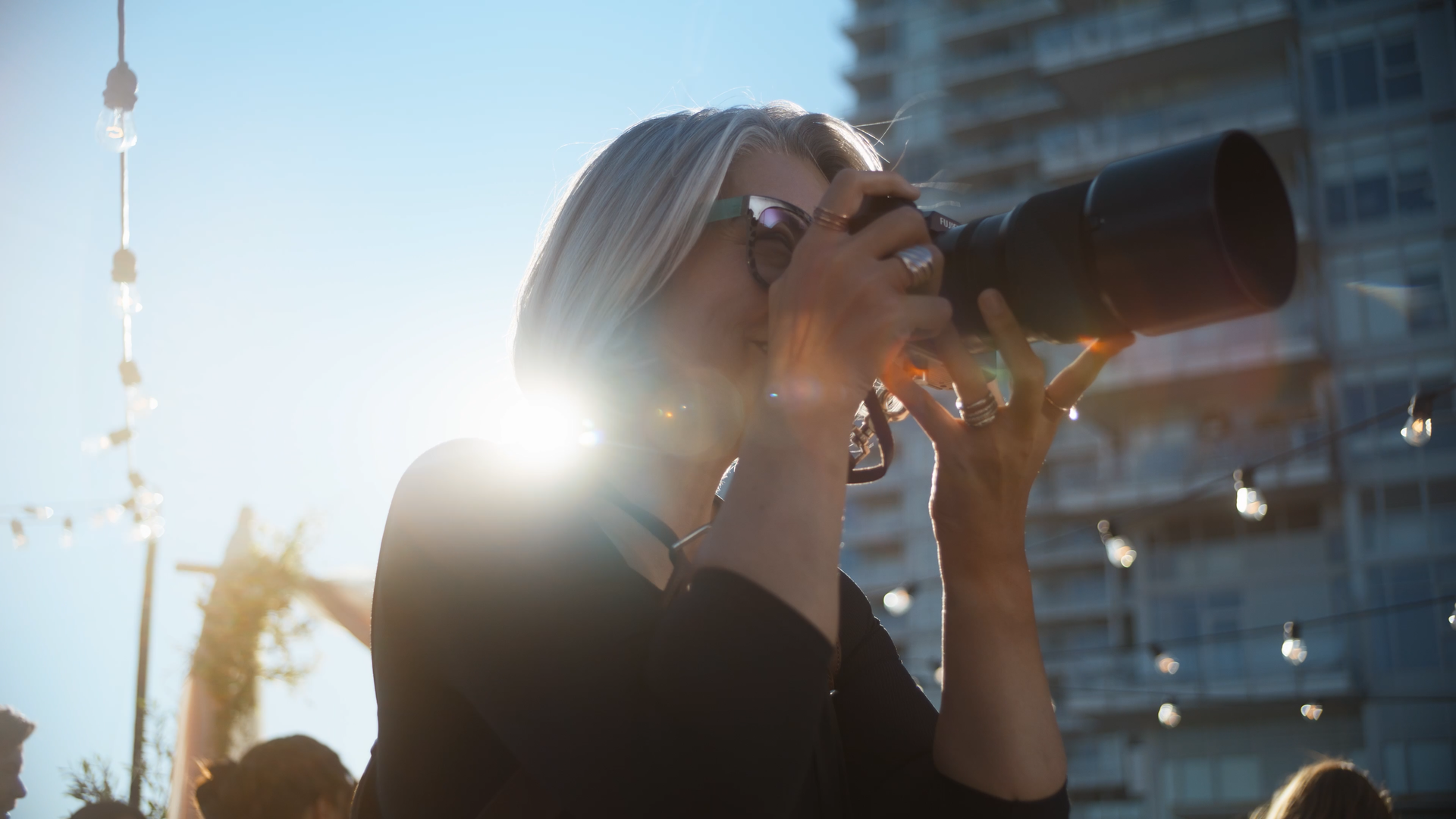 Woman with blonde hair and sunglasses holding a camera up to her face, taking a photo outdoors in bright sunlight.
