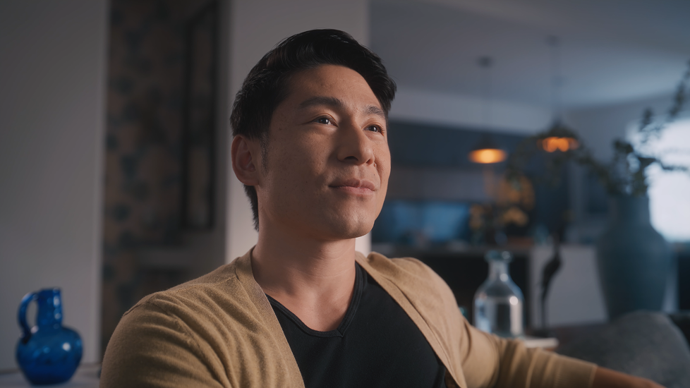 A young man sitting on a sofa in a modern living room, looking thoughtfully into the distance.