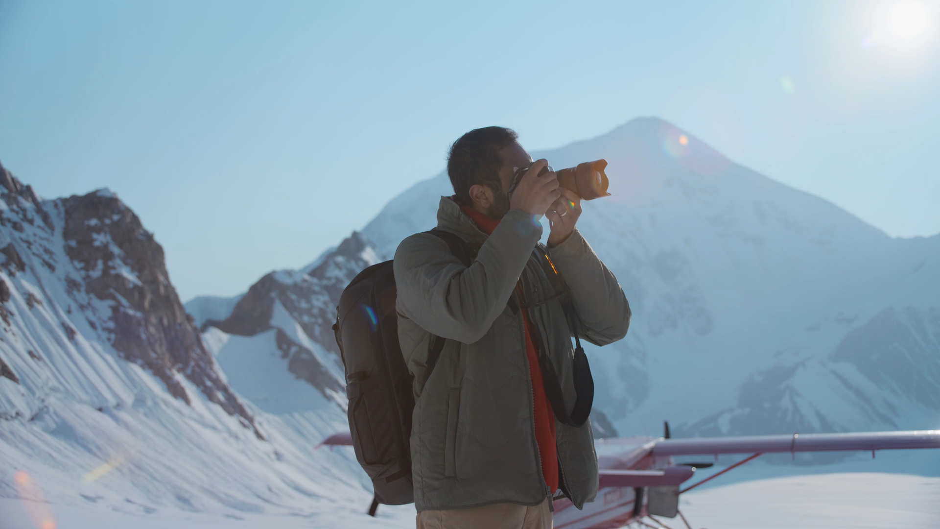 Man in winter gear taking a photo with a camera in snowy mountain landscape with a glacier and mountain peaks in the background.