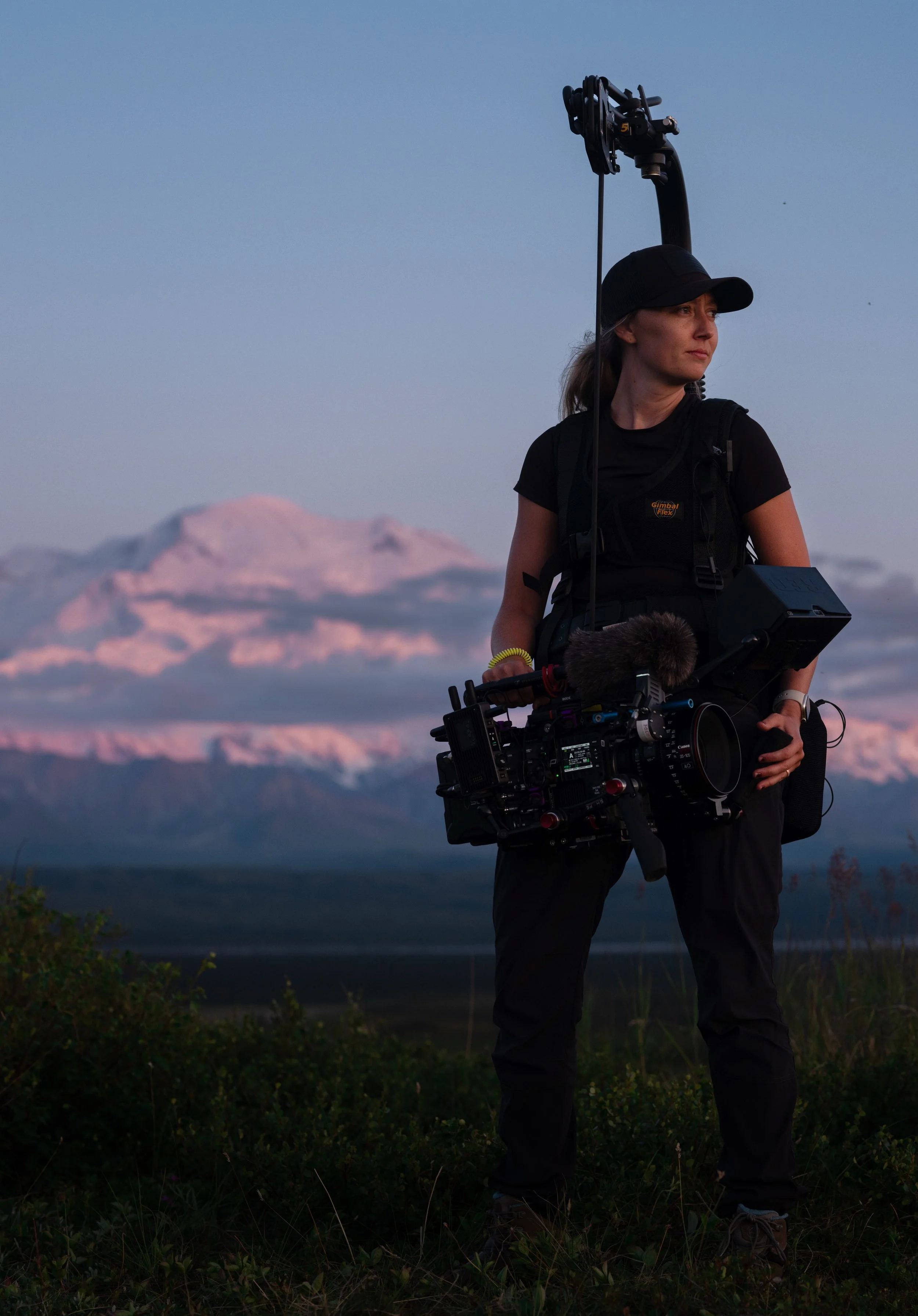 A woman wearing a black cap and black shirt is holding a professional video camera in an outdoor setting during sunset or sunrise. In the background, a snow-capped mountain is visible under a clear sky.