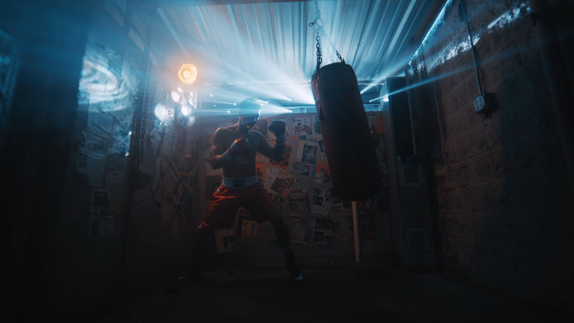 A boxer in red shorts practicing punches on a heavy bag inside a dimly lit gym with blue lighting and wall covered with newspapers.