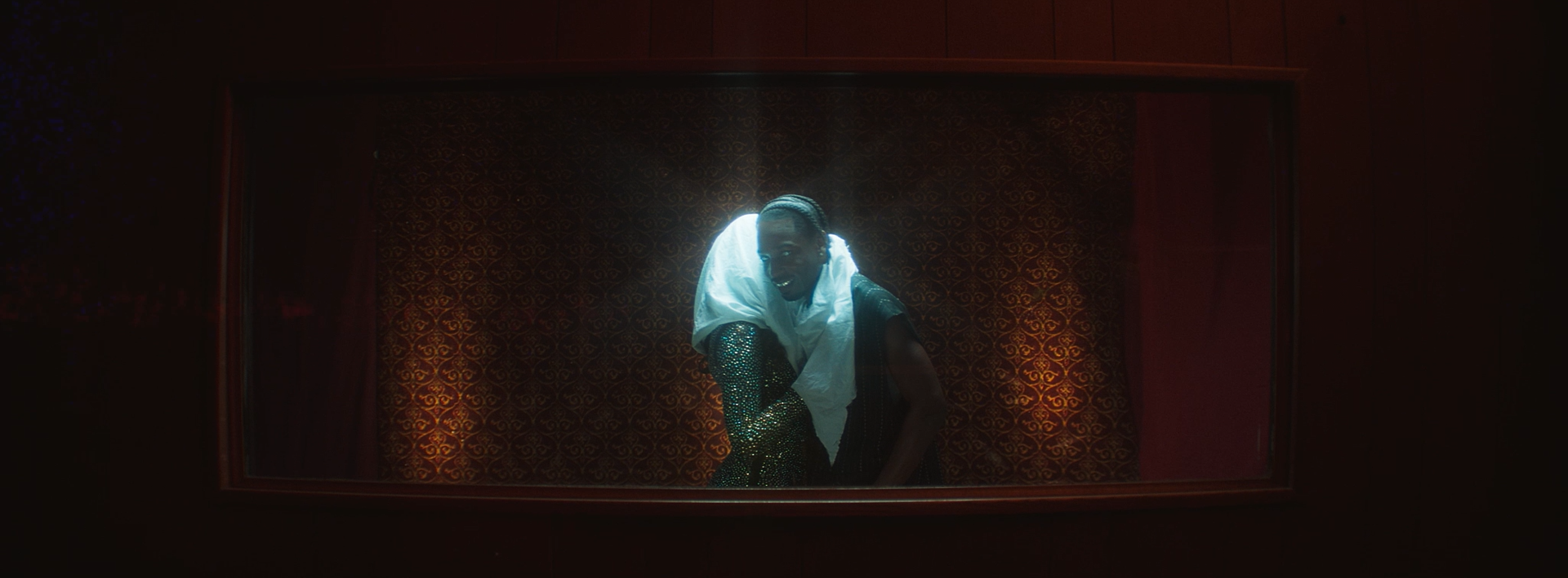 Person wearing a white shirt and shiny sequined pants standing behind a glass window with dim lighting.