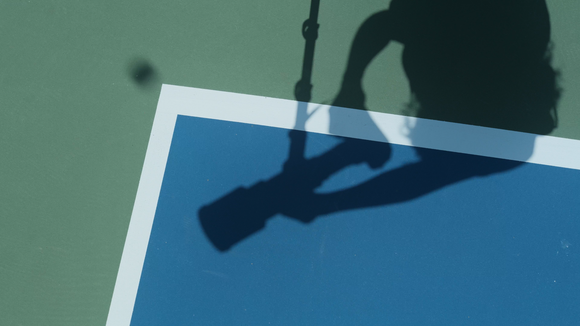 Shadow of a tennis player holding a racket on a tennis court with green and blue surfaces.