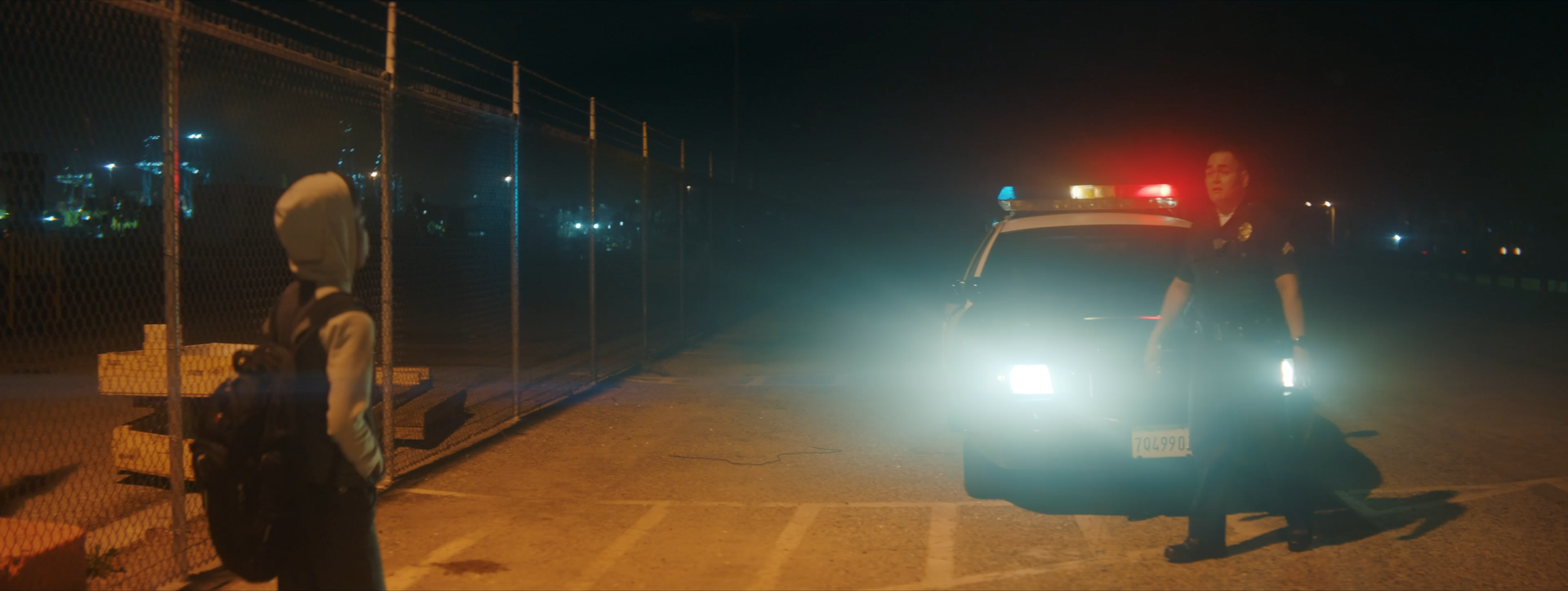 A police officer walking near a police car with flashing lights at night, a young person with a hoodie and backpack standing nearby.