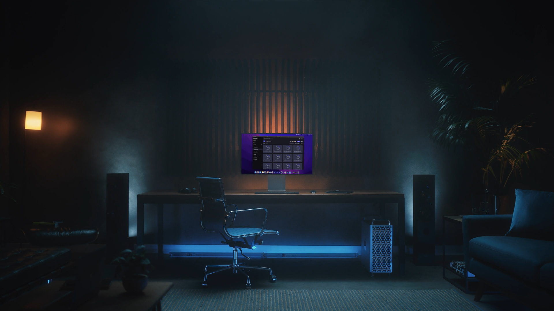 A dimly lit home office with a large monitor on a wooden desk, a black office chair, two tall speakers, and a potted plant on the right, illuminated by warm yellow and cool blue lighting.