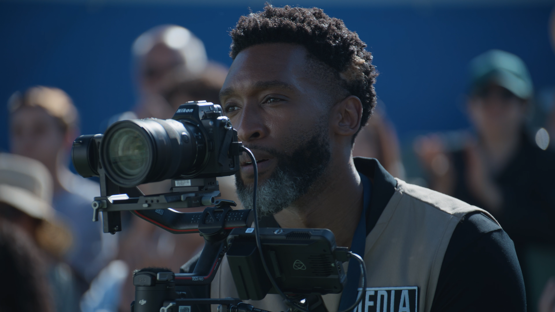A man operating a professional camera on a tripod at an outdoor event with a crowd in the background.