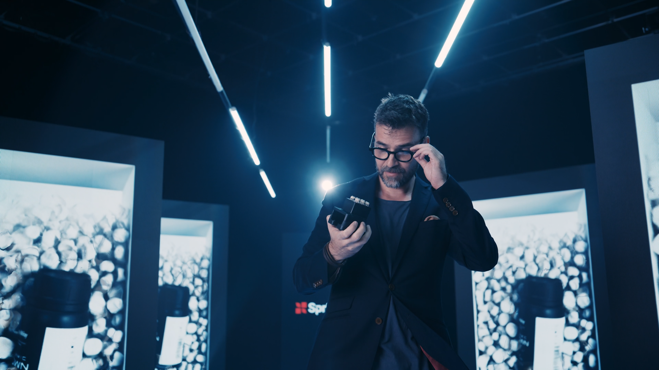 A man in glasses and a suit looks at a small device in his hand while standing in a futuristic room with blue lighting and large display screens.