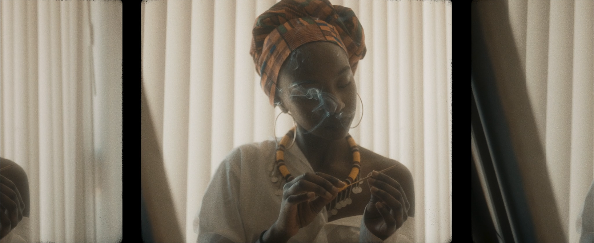 A woman wearing a colorful headwrap and large earrings, smoking and holding a necklace, standing in front of vertical blinds.