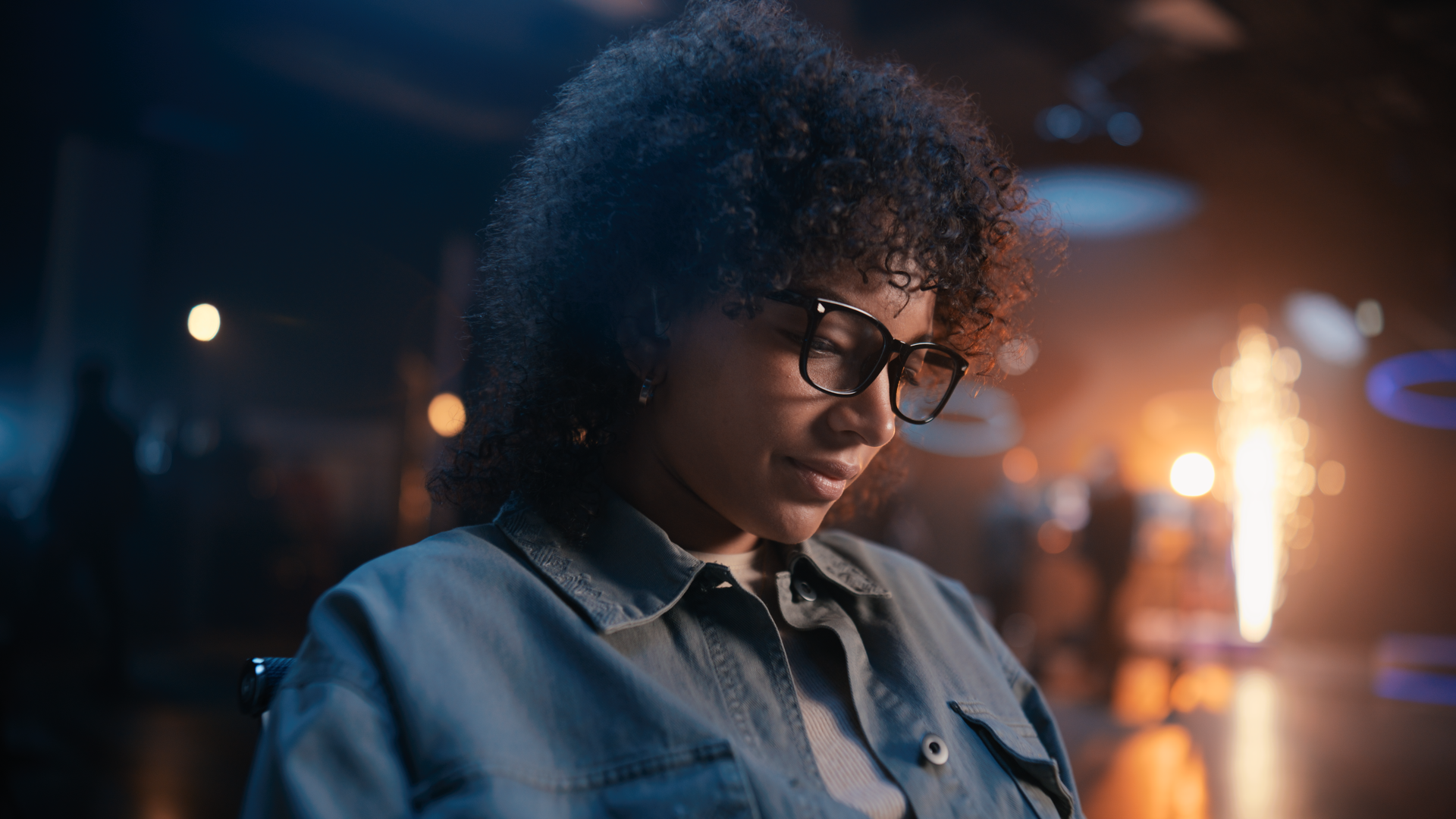 A young woman with curly hair and glasses, smiling and looking down, in a dimly lit indoor setting with blurred lights and a festive atmosphere.
