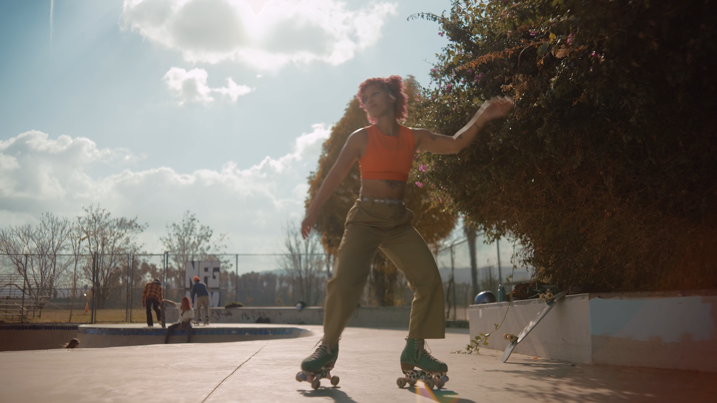 A young woman with pink hair rollerblading at a skate park during daytime, with people in the background and cloudy sky.
