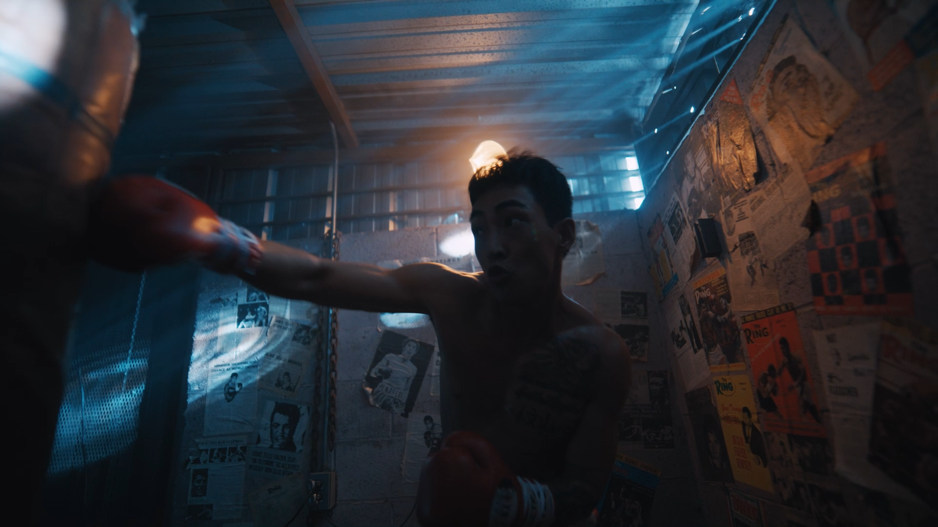 A young male boxer training in an underground boxing gym with newspaper-covered walls, wearing red boxing gloves and throwing a punch.