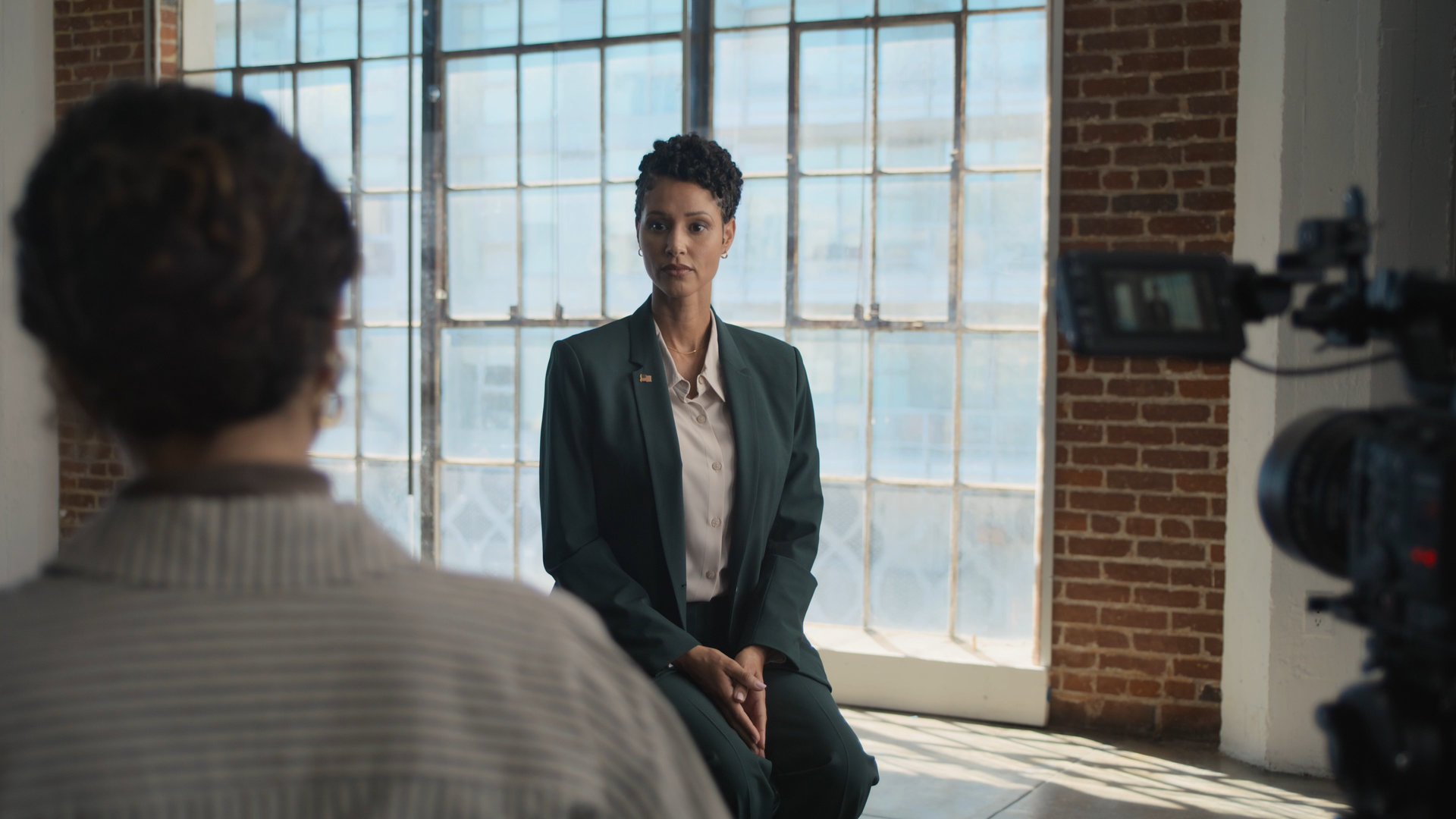 A woman in a dark green blazer and white blouse sitting in front of a large industrial-style window during an interview or filmed conversation, with a camera crew capturing her on video.