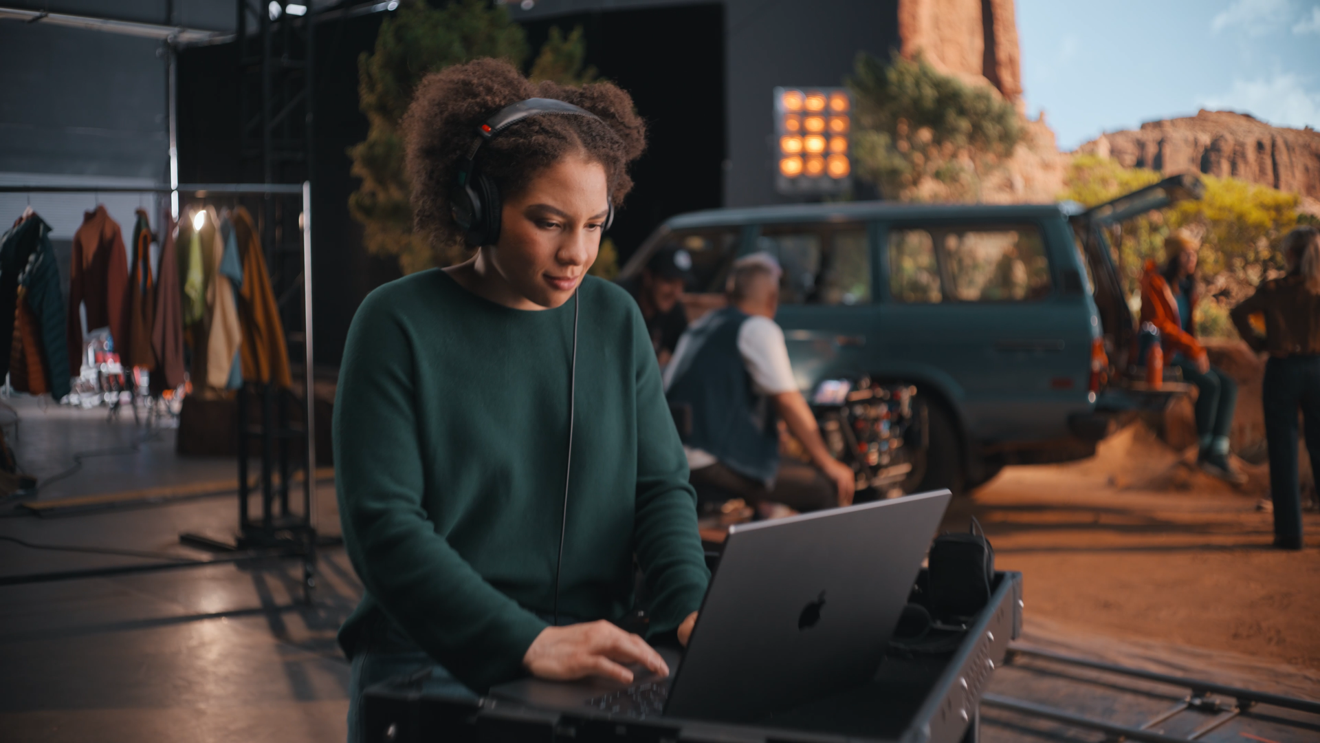 A woman working on a laptop with headphones on, on a film or TV set outdoors, with crew members, equipment, and a vehicle in the background amidst a desert landscape.
