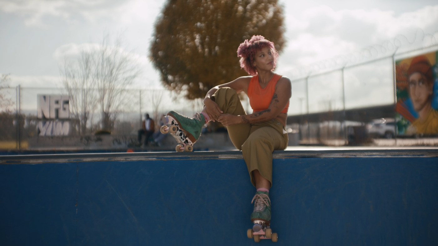 Young woman with pink curly hair sitting on the edge of a skate park bowl, preparing to skate, with a large tree and graffiti in the background.