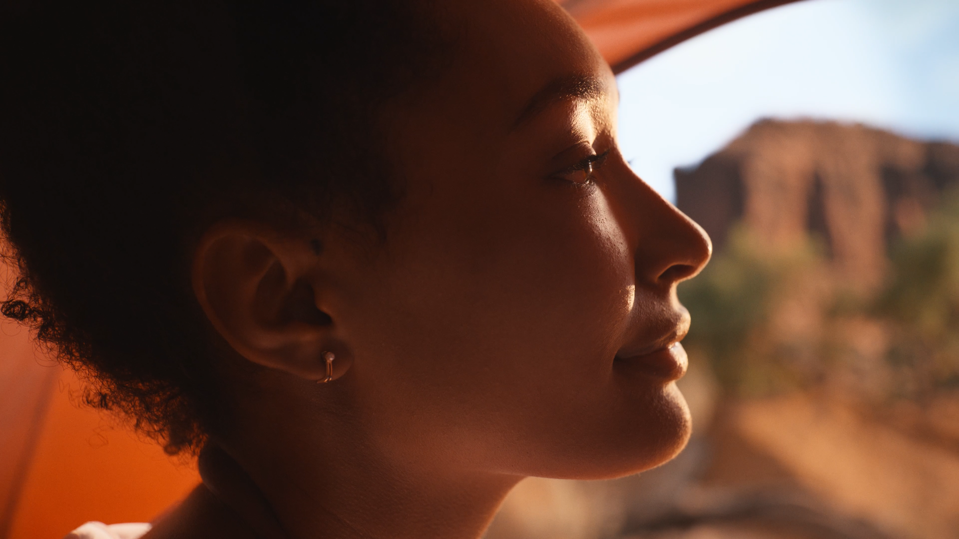 Close-up profile of a young woman with short curly hair, wearing a small hoop earring, gazing into the distance with a desert landscape in the background during sunset.