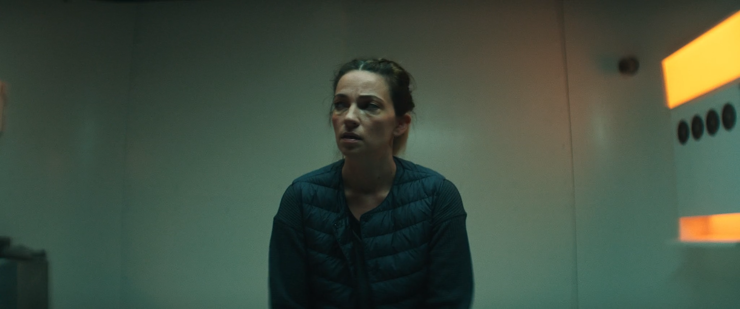 A woman sitting in an elevator with a distressed expression, wearing a dark quilted jacket, with orange lights on the elevator walls.