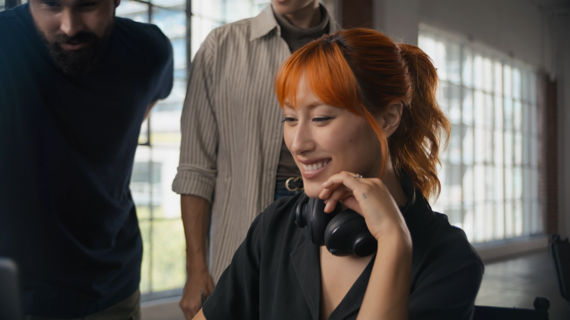 A woman with orange hair smiling, wearing a black shirt and headphones around her neck, looking at a device. Two people, a man with a beard and a woman, leaning in and looking at the same device, in an indoor space with large windows.