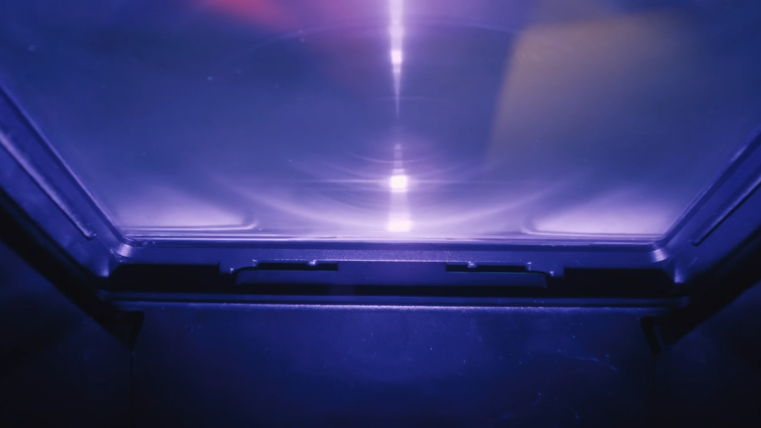 Close-up of a metallic, reflective surface illuminated by purple and blue lights, with a directional light source at the top center creating a bright streak and circular reflections.