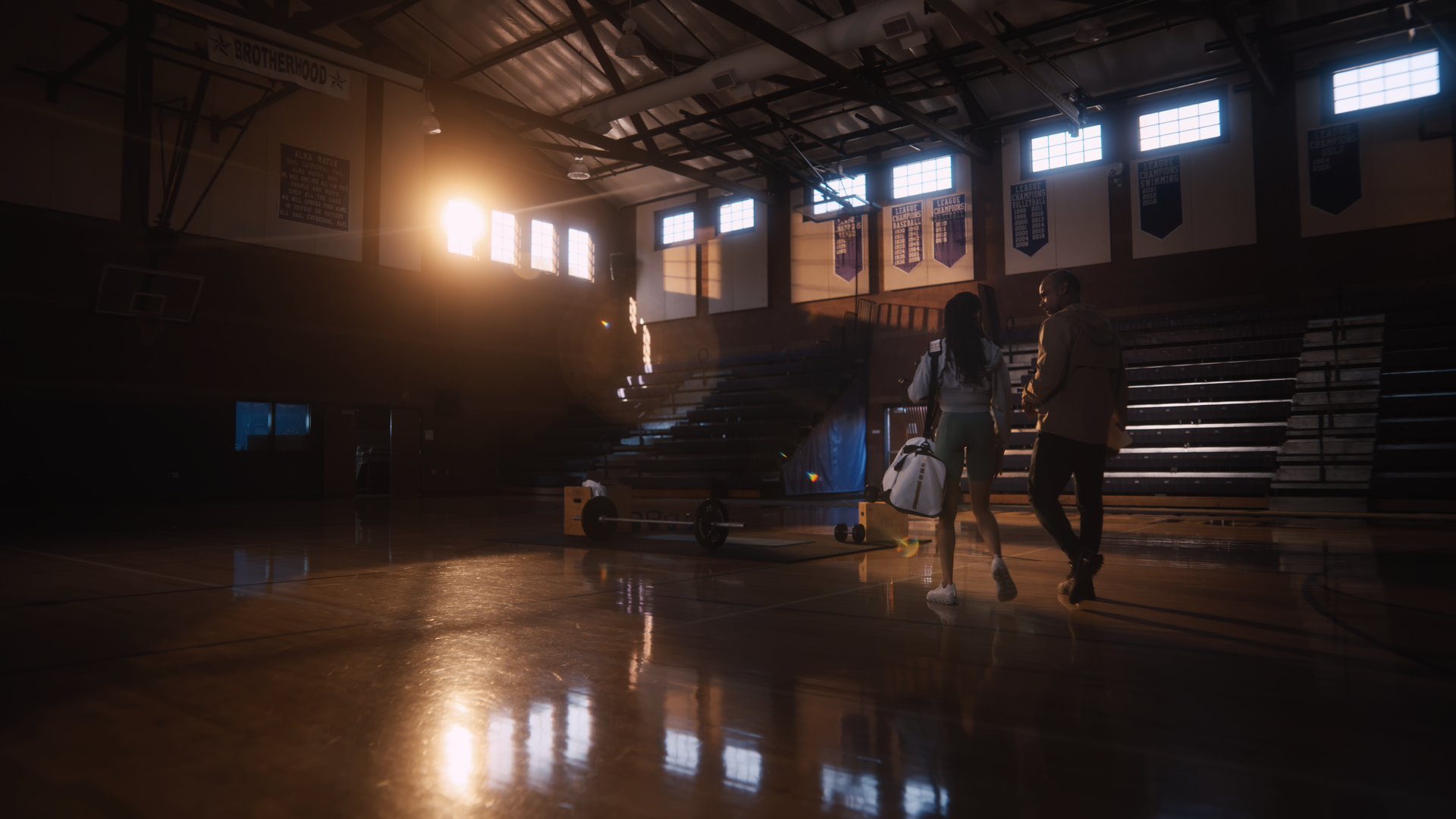 Two people walking into a gymnasium with gym equipment, sunlight streaming through large windows, and banners hanging on the walls.
