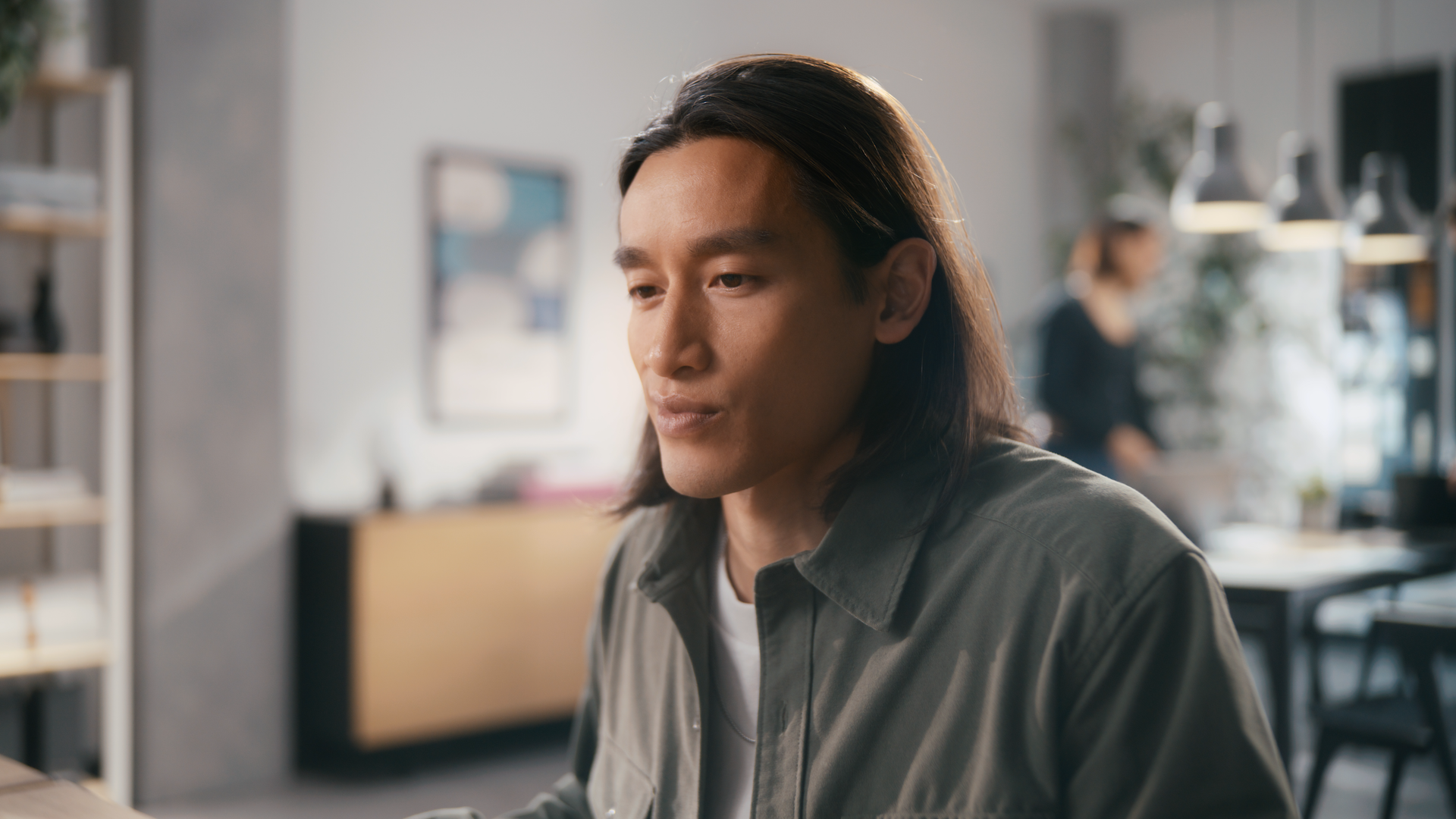 A young man with long dark hair sitting at a table in a modern office or cafe, looking down thoughtfully with blurred background of another person in the distance.