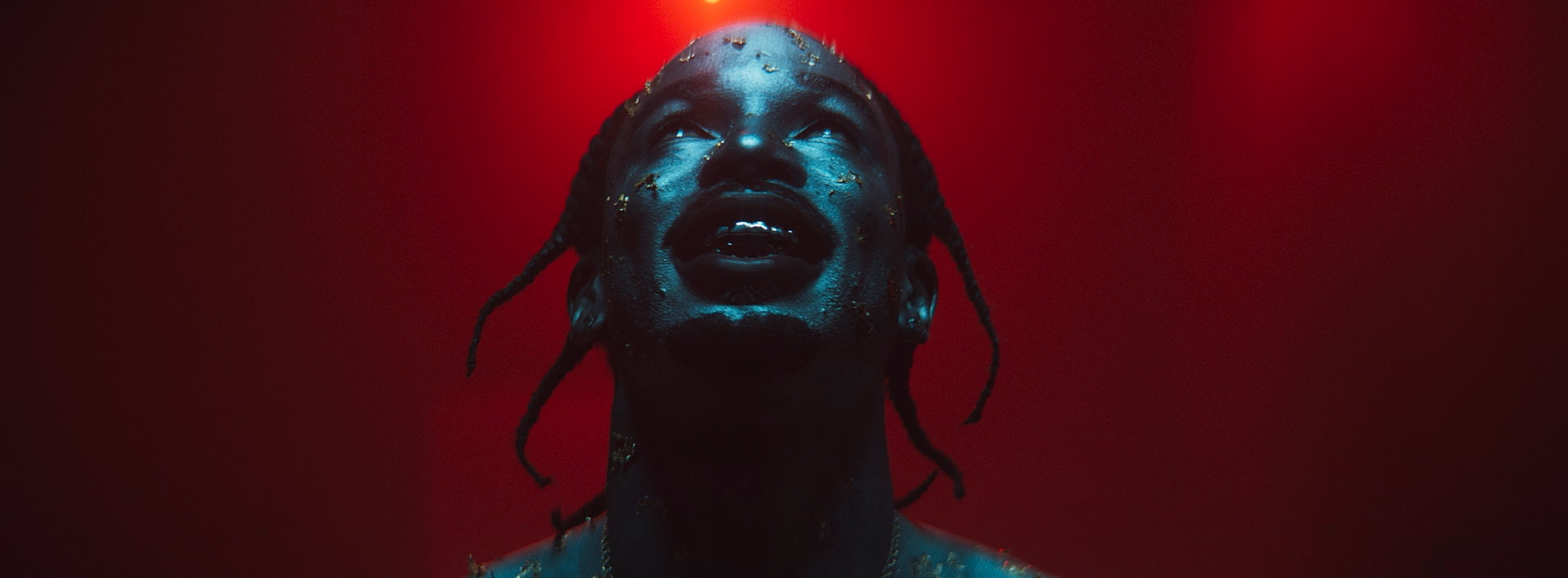 Close-up of a person with dark skin and braided hair, looking upward with a joyful expression, illuminated by red light.
