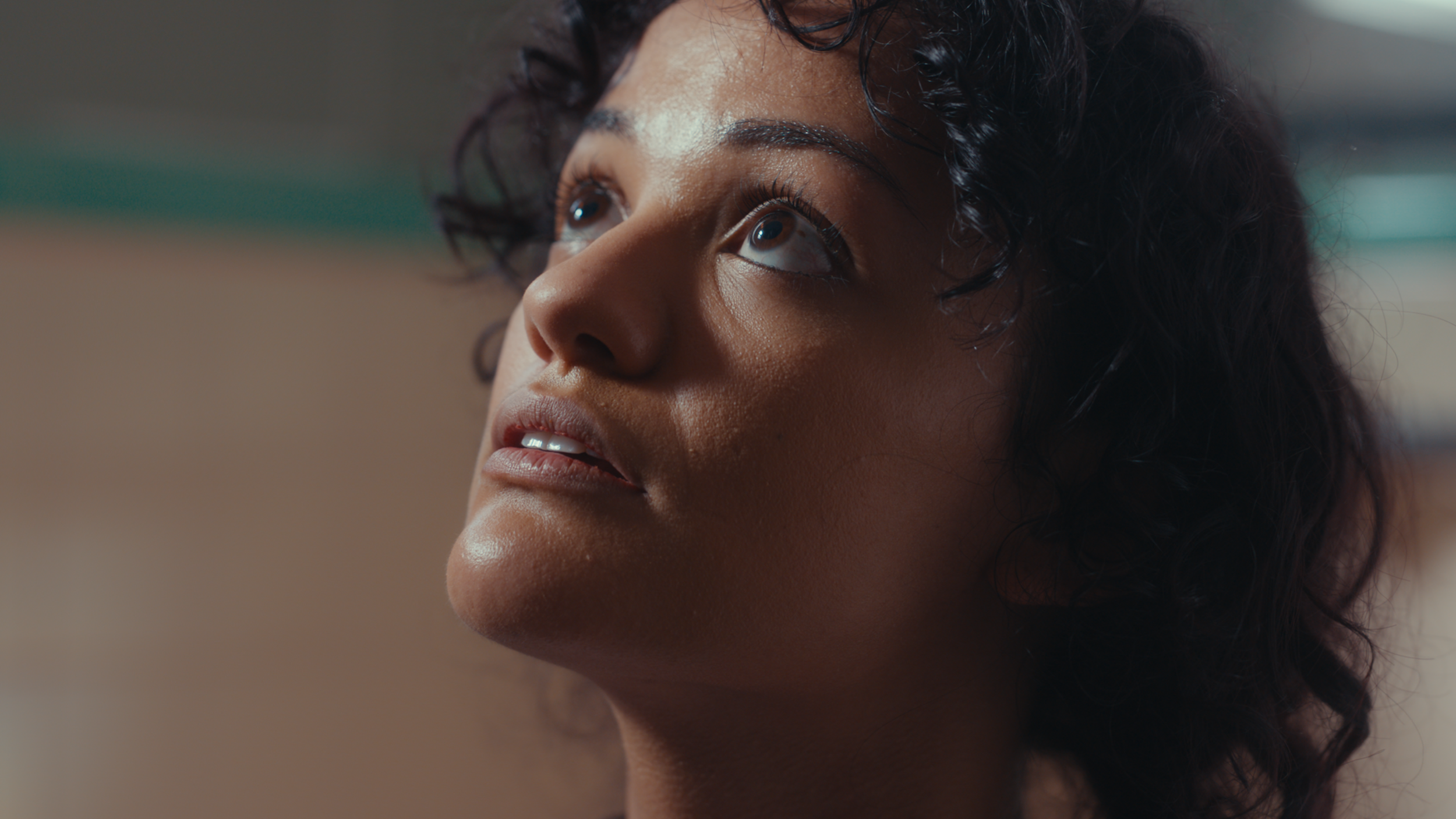 Close-up of a woman with dark curly hair looking upwards, with natural lighting highlighting her face and skin texture.