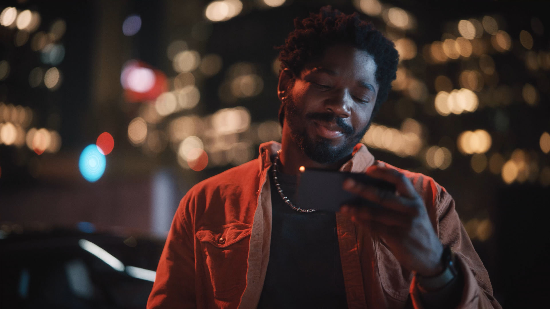 A man with curly hair and a beard smiling and looking at his phone at night, with blurred city lights in the background.