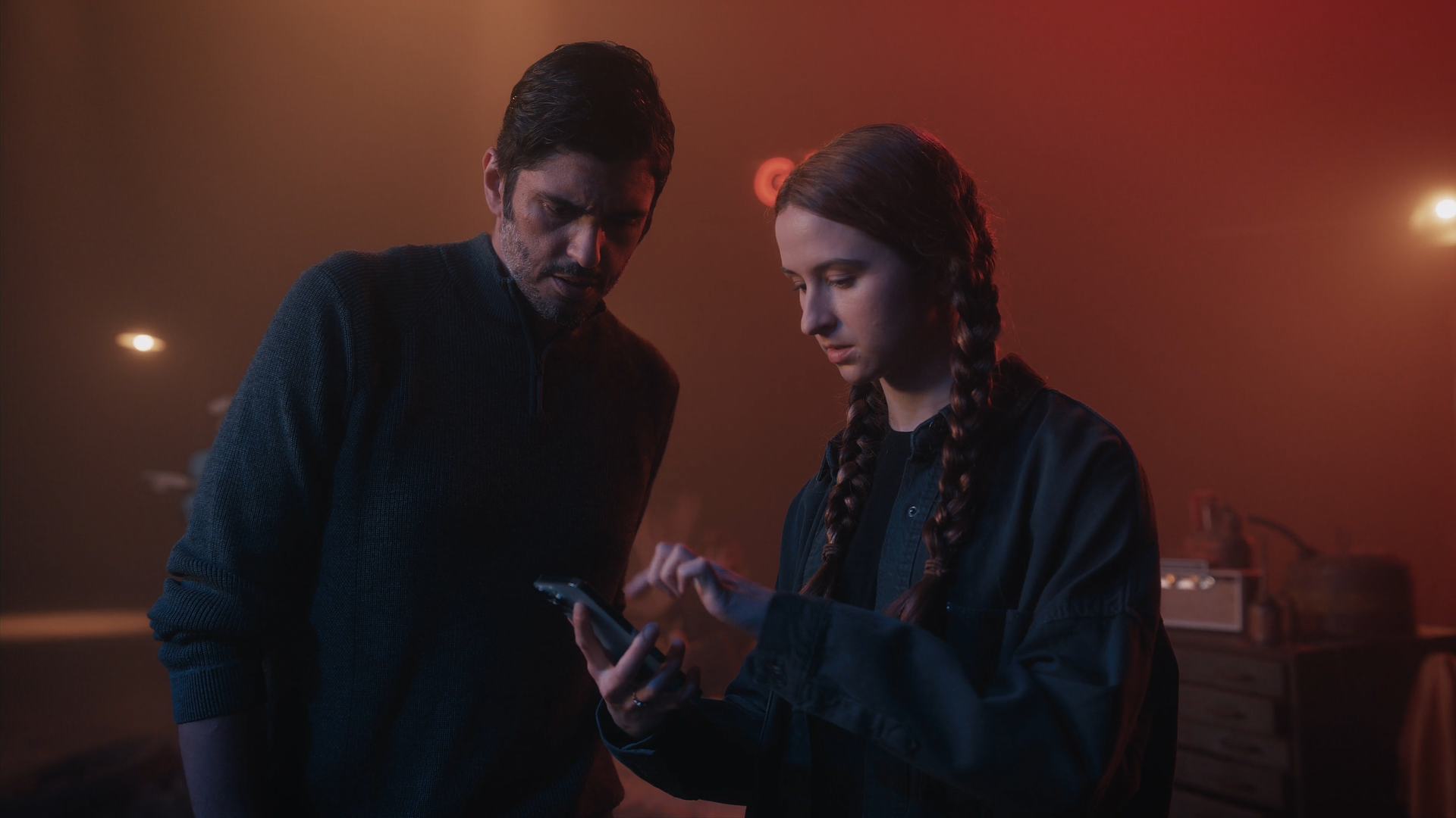 A man and a woman with braided hair look at a smartphone together in a dimly lit room with red lighting.
