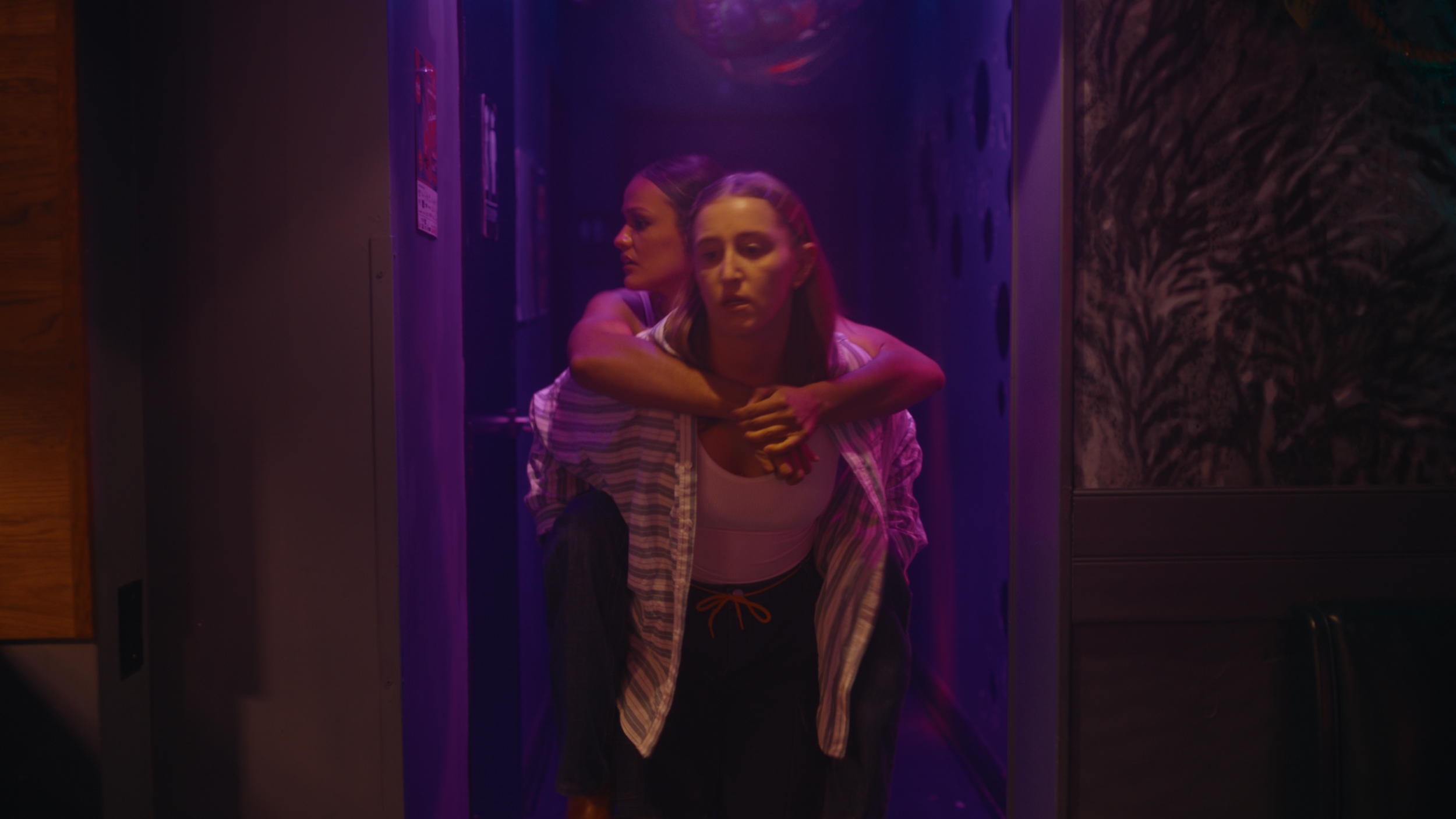 Two women are in a dimly lit hallway. One woman is giving the other a piggyback ride, and both appear to have serious or contemplative expressions. The lighting is primarily purple and pink.