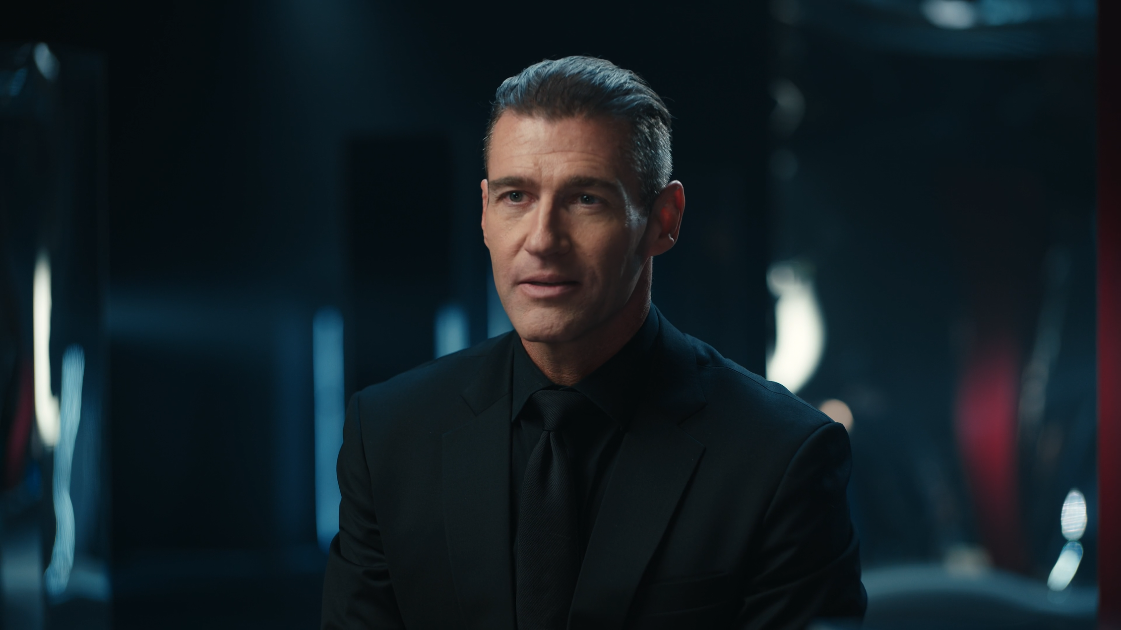 A man in a black suit with slicked-back hair looking to the side in a dark, modern setting with blurred lights in the background.