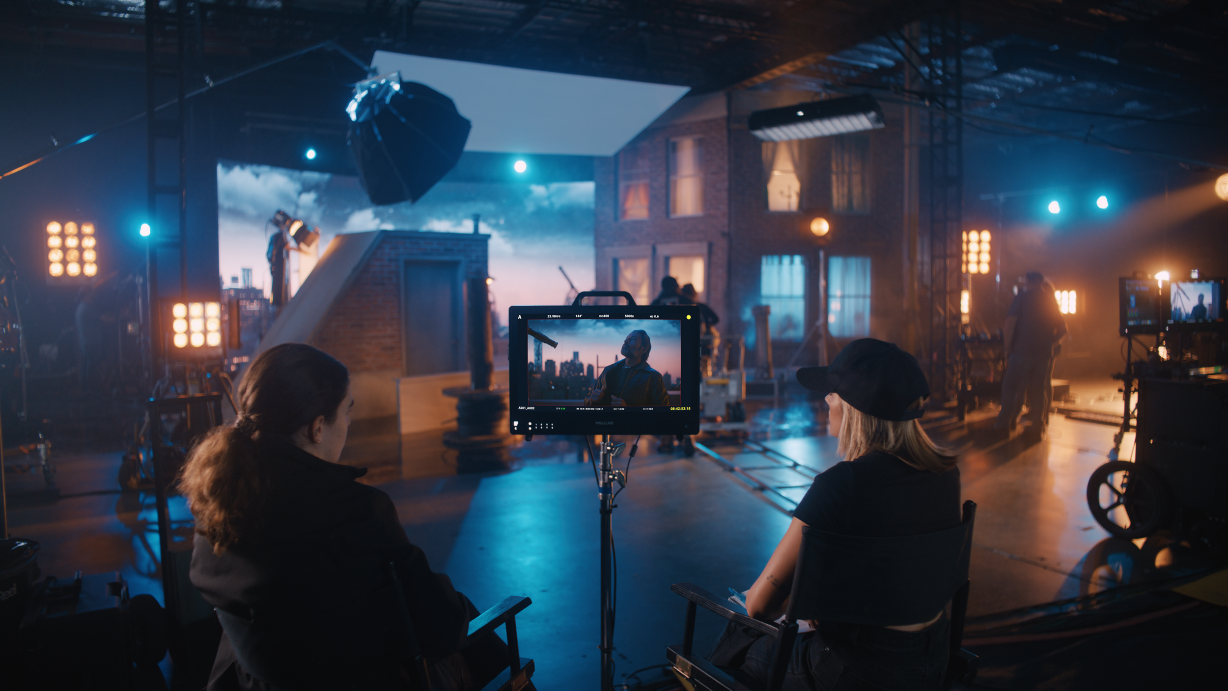 Film crew on set during shot, with director viewing scene on monitor, lighting equipment, and a large greenhouse-style window in the background.