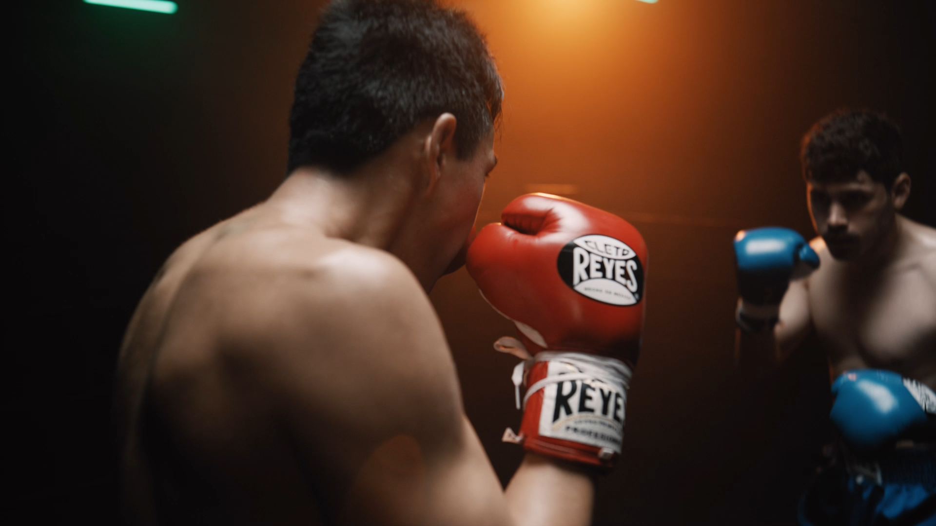 Two men in a boxing ring facing each other; one man in red gloves and the other in blue gloves, with a dark background and warm lighting.