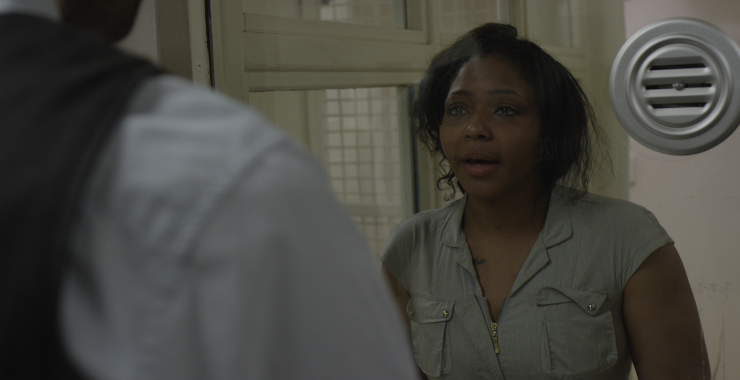 A woman with dark, curly hair and a light-colored shirt appears to be talking to a person whose back is facing the camera, in an indoor setting near a window or glass door.