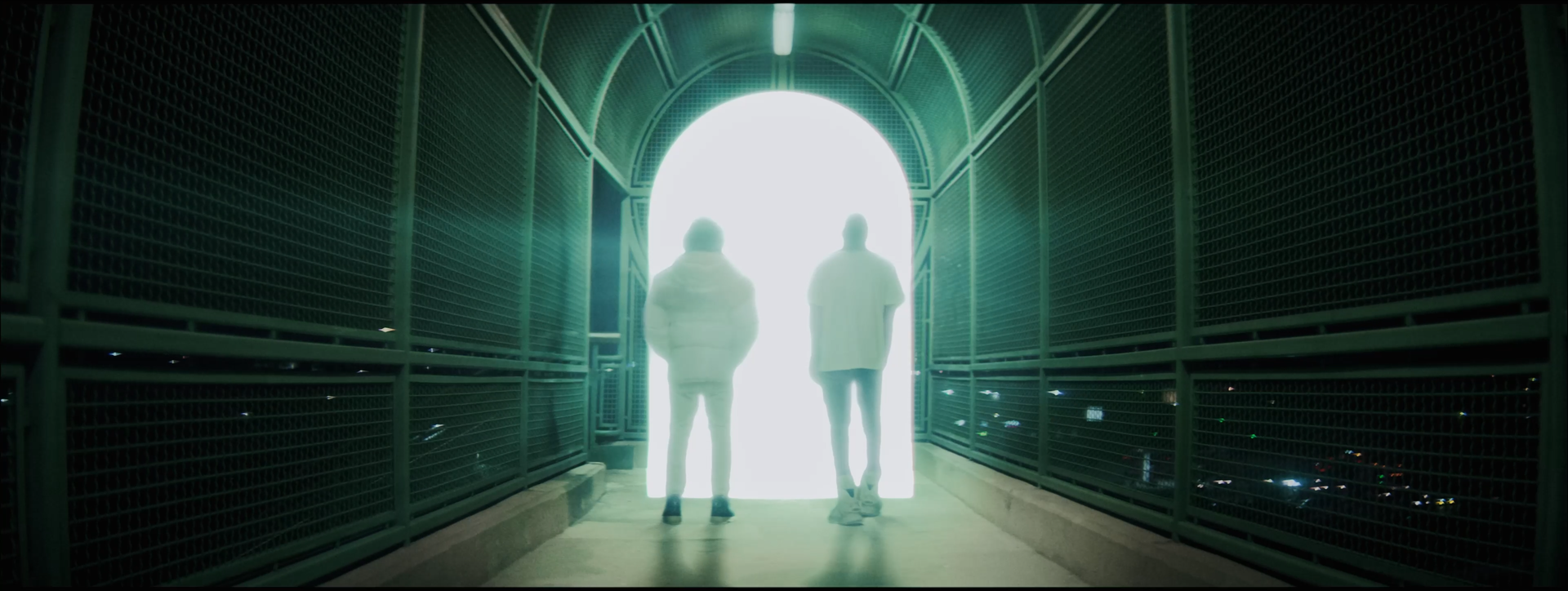 Two people walking through a dark, enclosed pedestrian bridge towards a bright, overexposed exit.