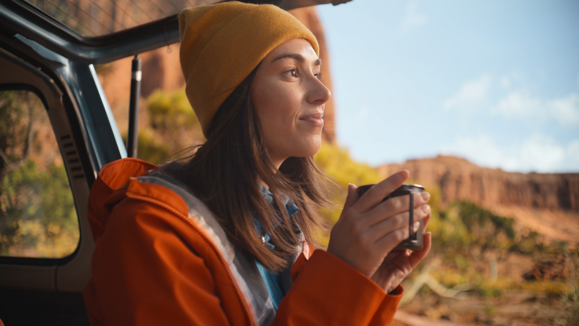 A woman with brown hair wearing a yellow beanie and orange jacket sitting in a vehicle, holding a mug, with desert canyon landscape in the background.