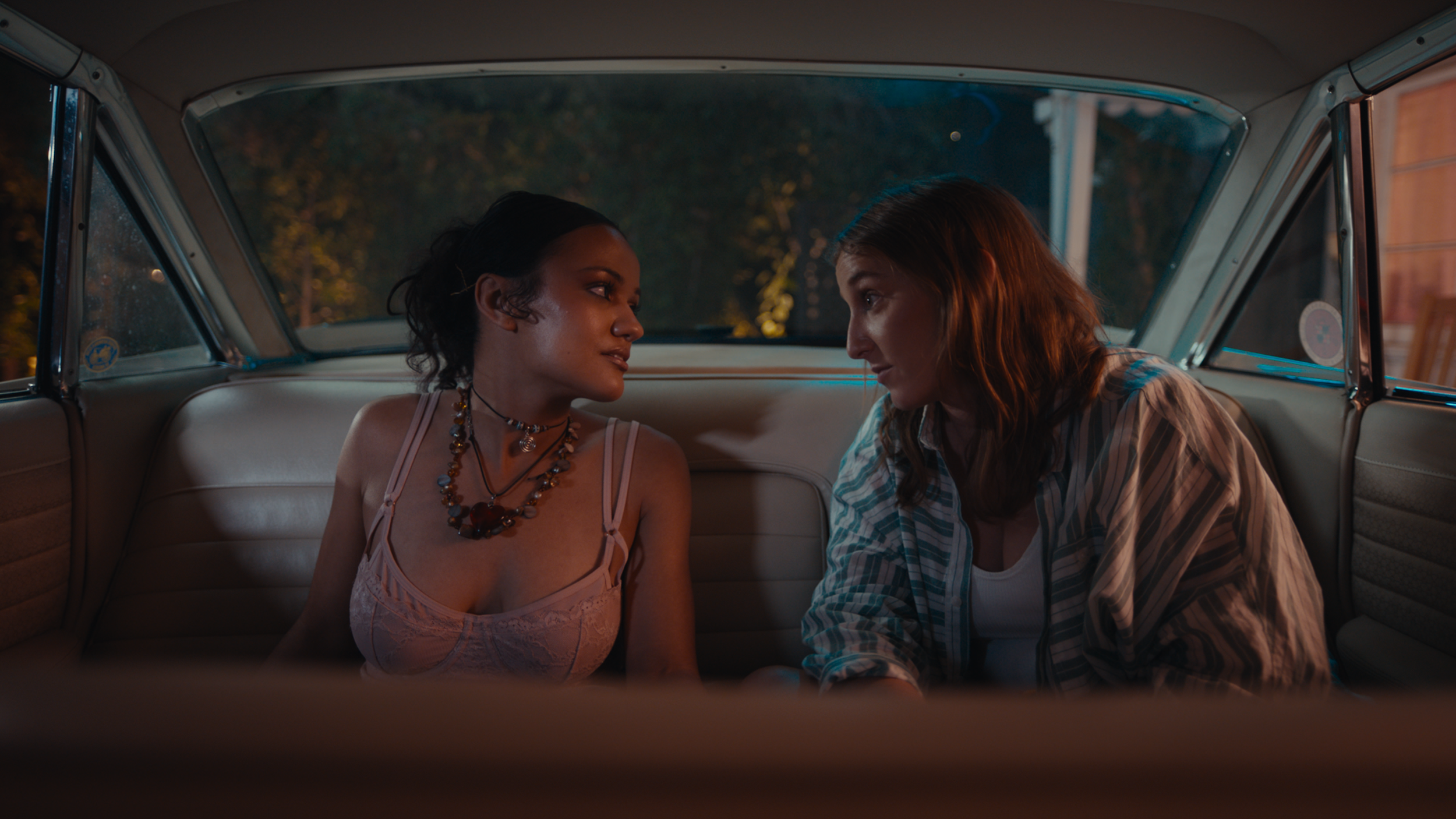 Two women sitting close together inside a vintage car at night, facing each other, with trees and city lights visible through the window.