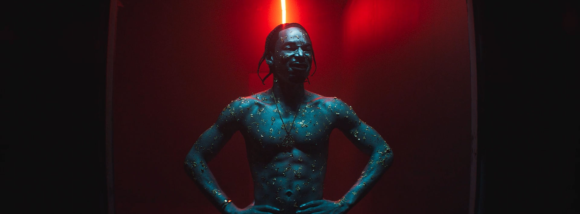 A shirtless man covered in gold leaf from the waist up, standing with hands on hips in a dark room with red lighting.