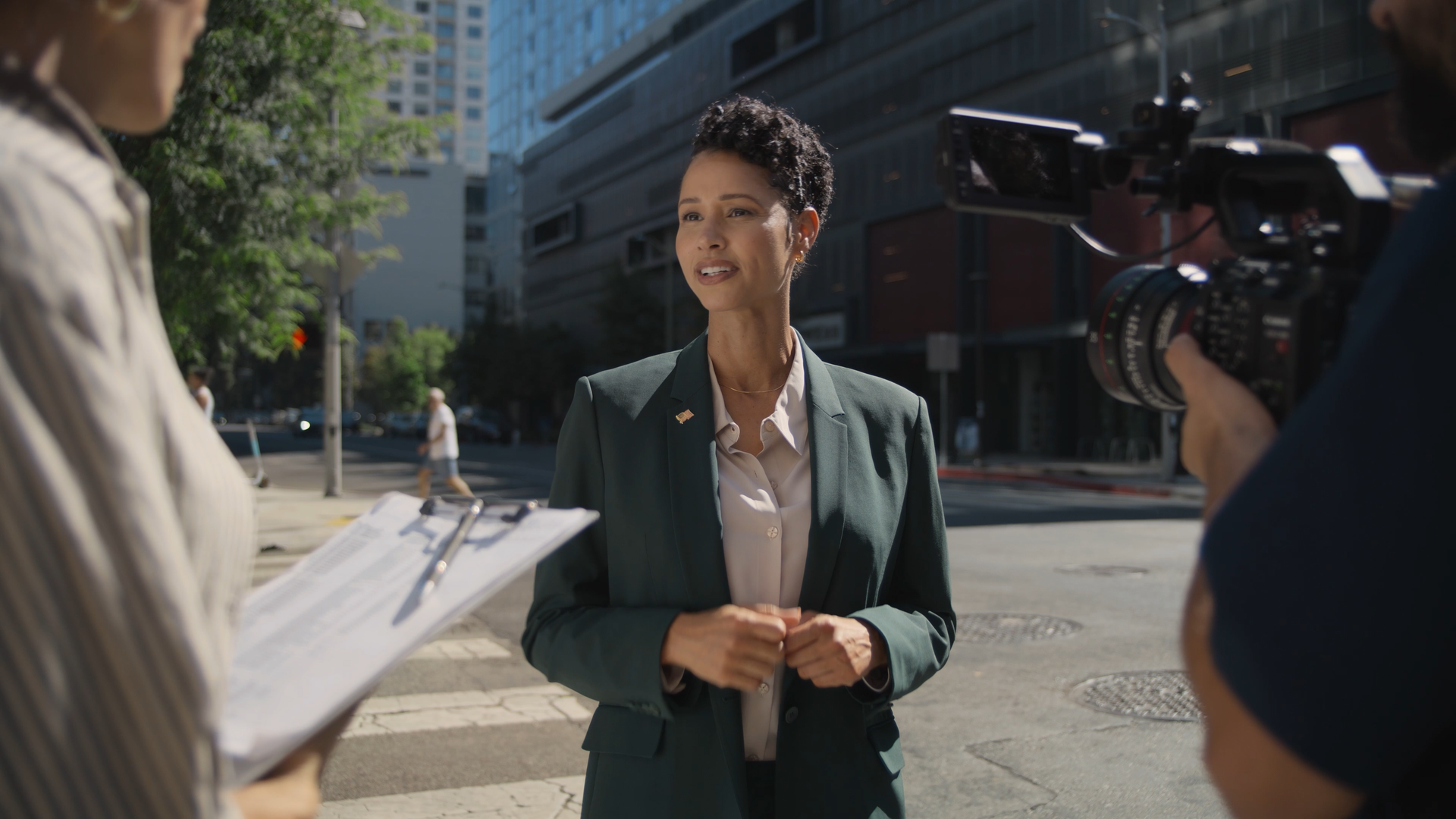 A woman in a green blazer being interviewed outdoors on a city street, with a camera operator recording her. A person with a clipboard is partially visible on the left.