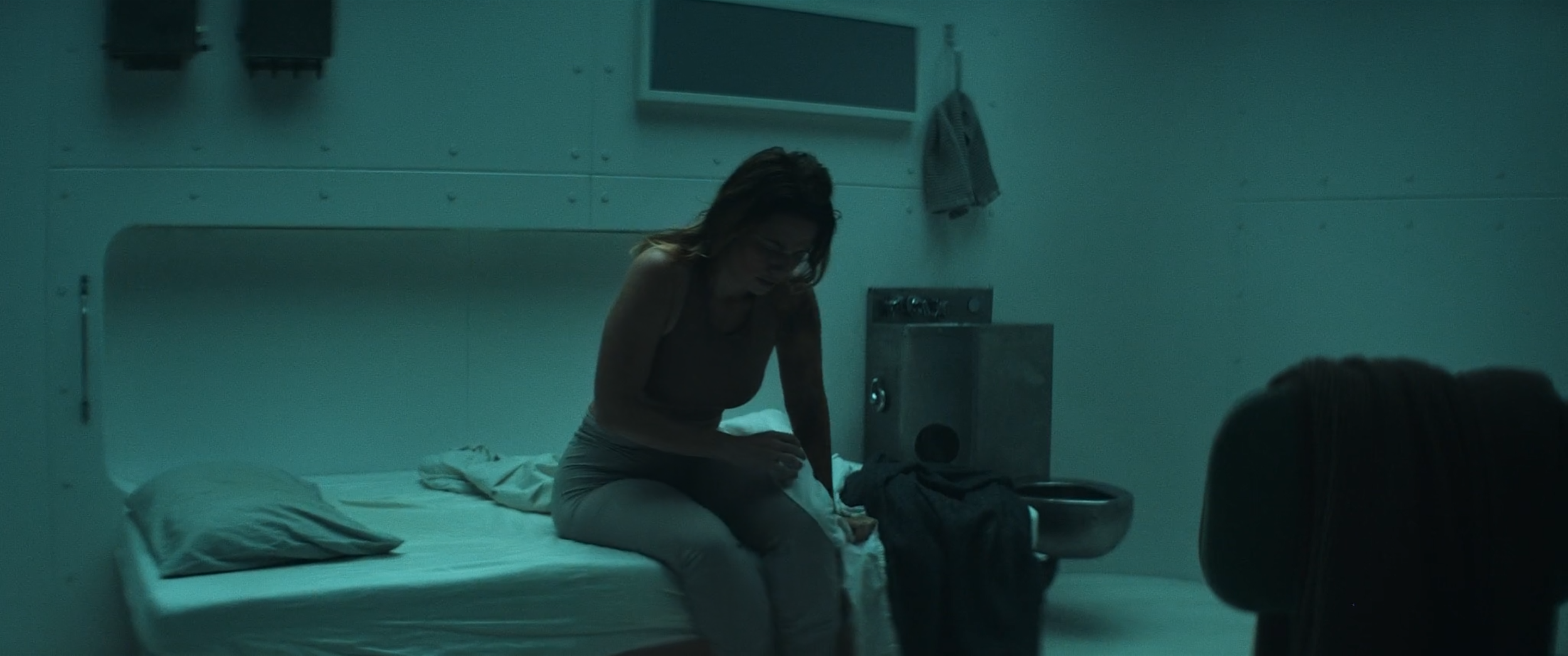 A woman with glasses sits on a hospital bed in a dimly lit room, looking down and touching her leg, with an open wastebasket nearby.