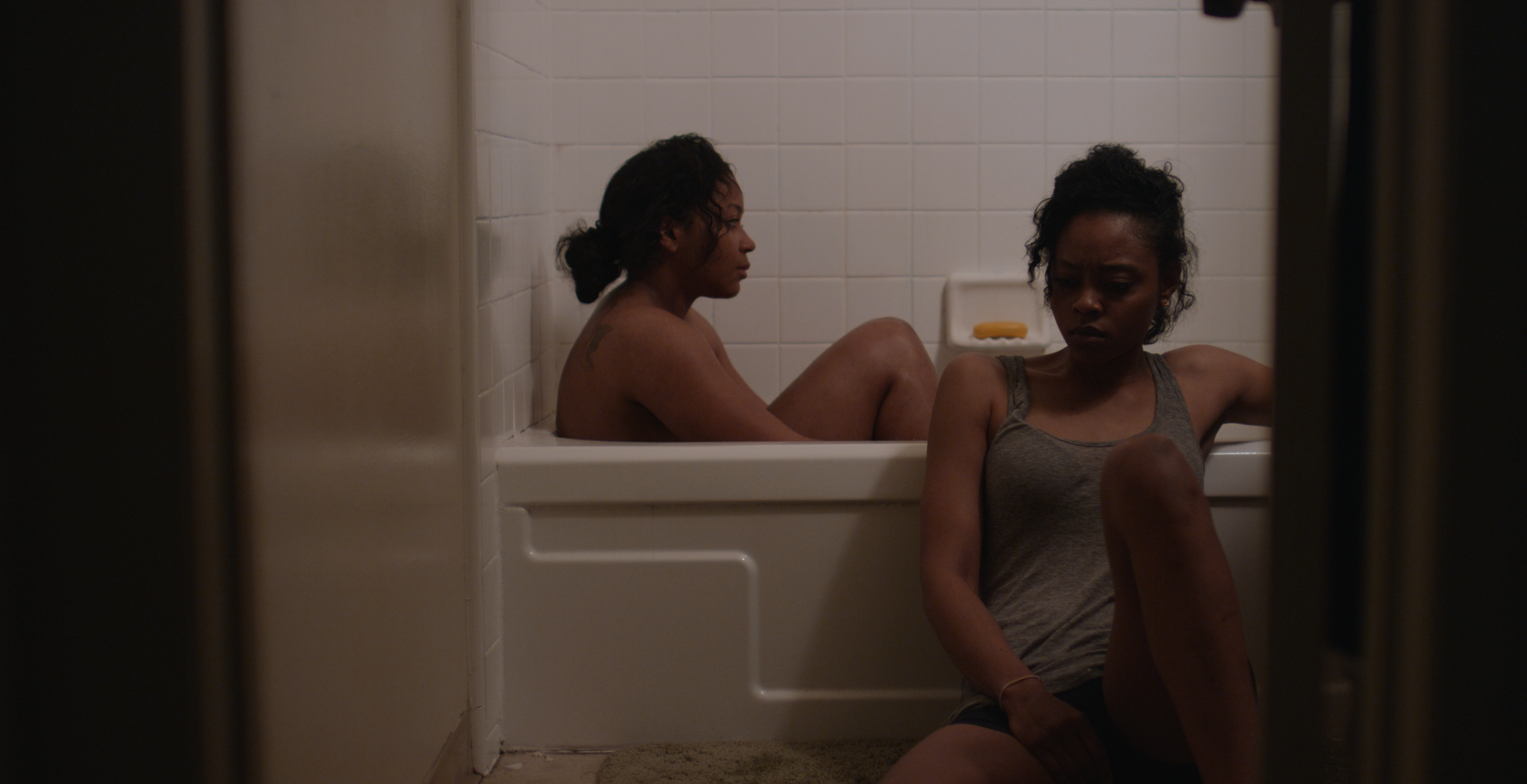 Two women sitting in a bathtub in a tiled bathroom, one with curly black hair sitting with knees up, and the other in a gray tank top, with a bar of soap on a soap dish behind them.