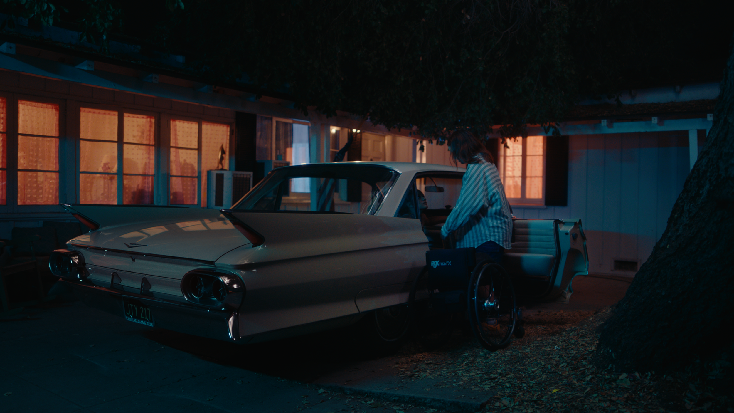 A woman with brown hair in a plaid shirt standing next to a vintage white car parked in front of a house at night. The house has lit windows with curtains, and there's a large tree in the foreground. The scene has a warm, nighttime glow.
