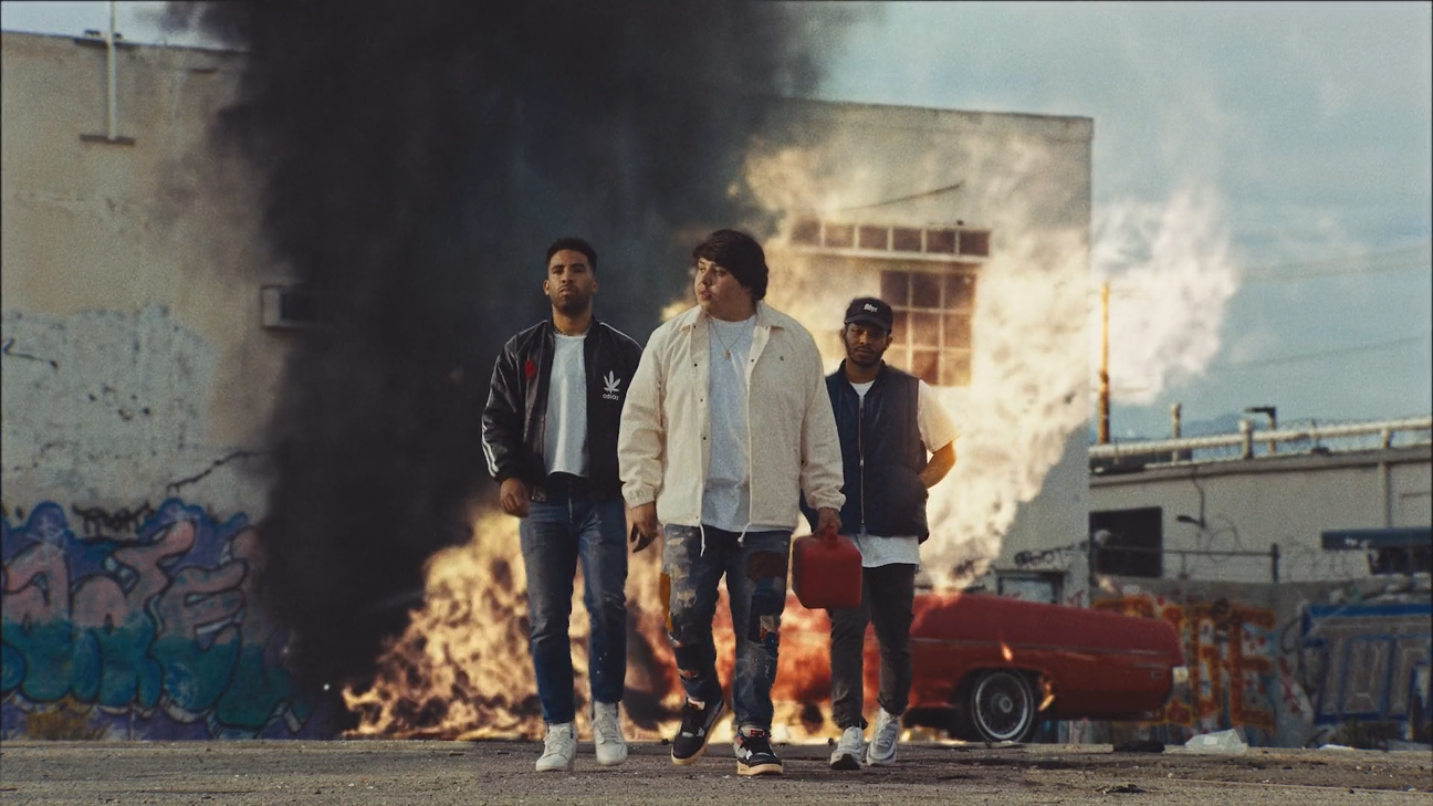 Three young men walk confidently in front of a burning building with thick black smoke. The scene appears urban with graffiti on the walls and a red car in the background.