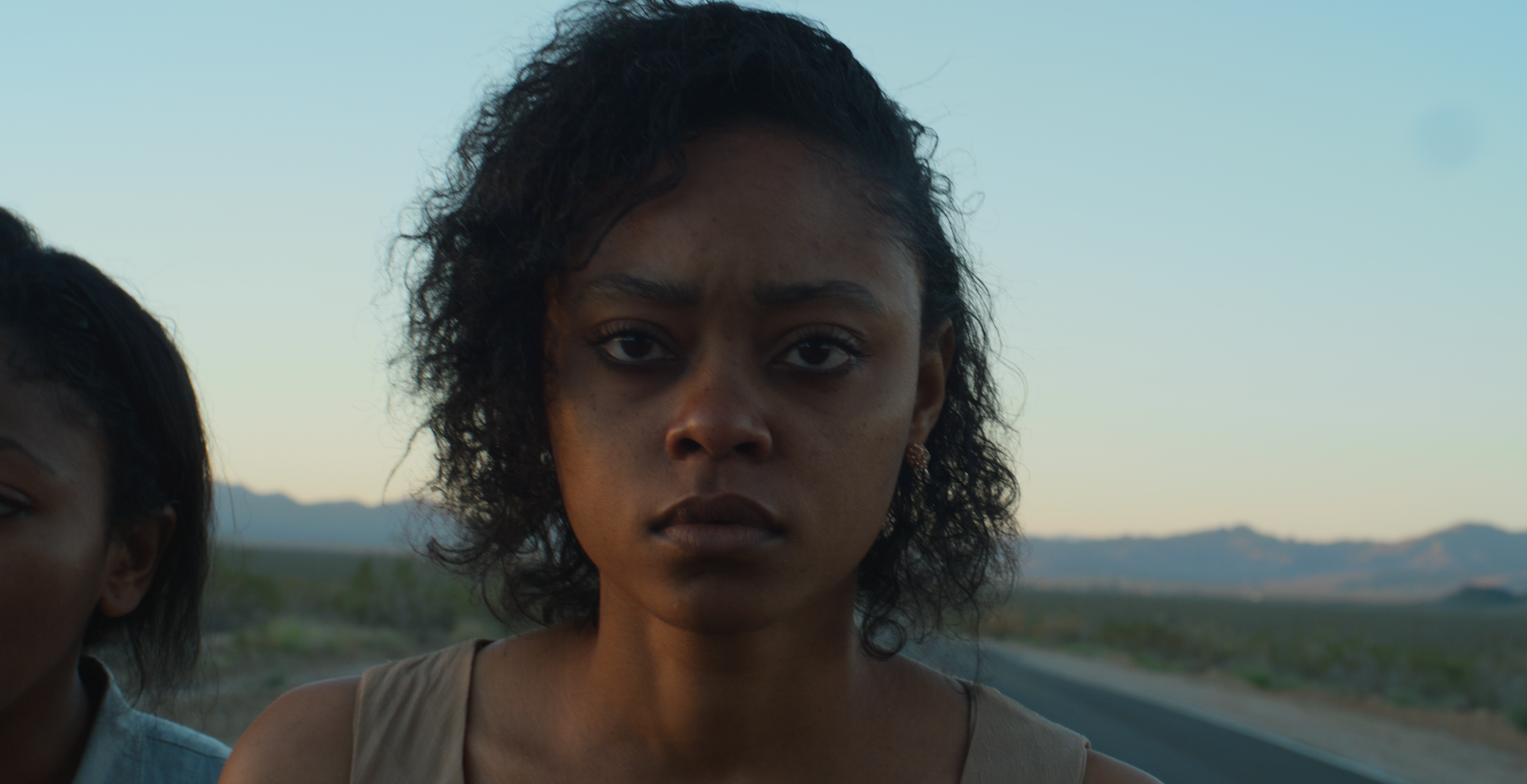 A woman with dark curly hair looking directly at the camera, with a serious expression, standing outdoors during sunset with mountains and a clear sky in the background.