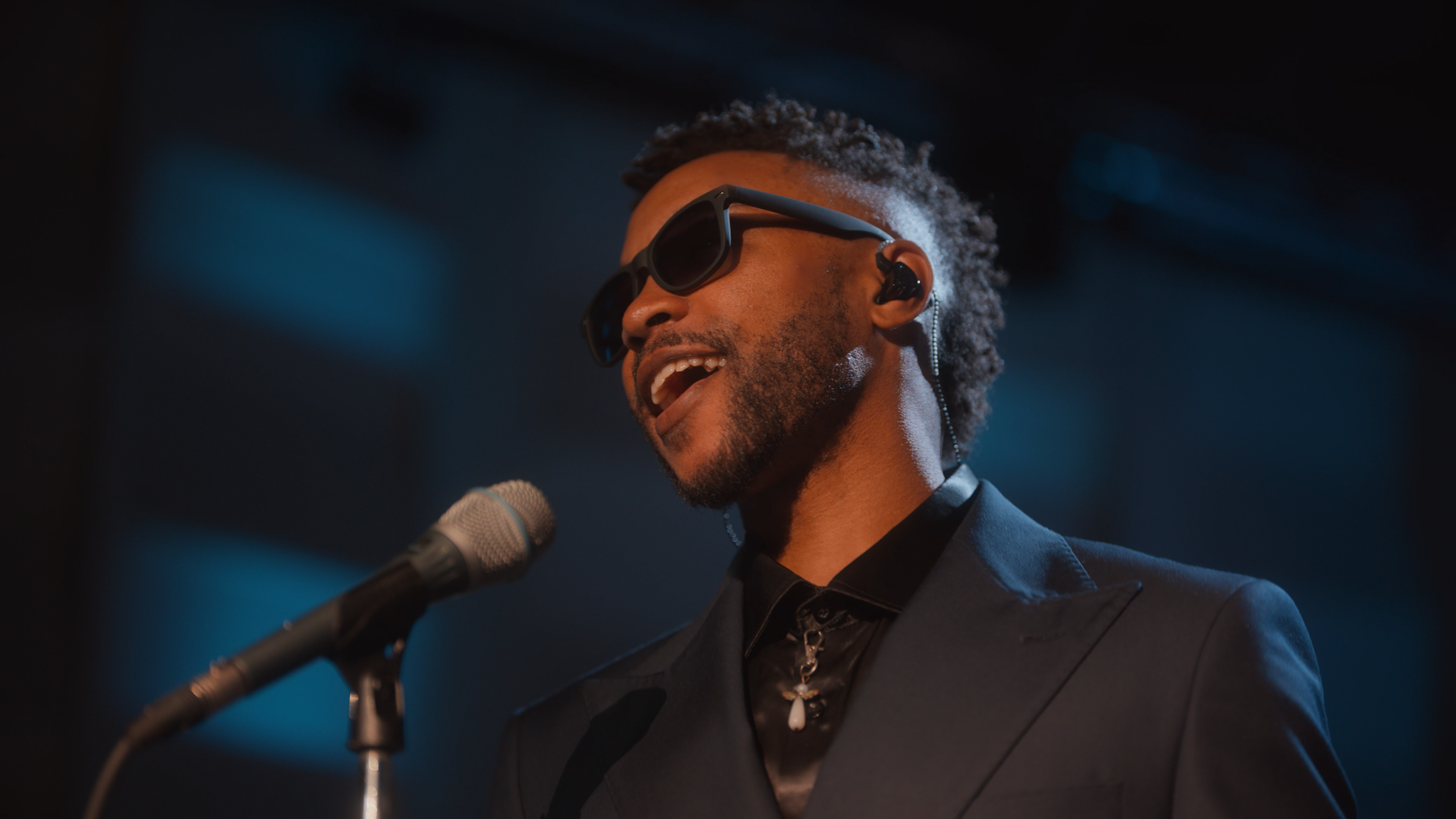 A man with curly hair and sunglasses singing or speaking into a microphone on stage, wearing a dark suit and a decorative necklace, with ear monitors visible.
