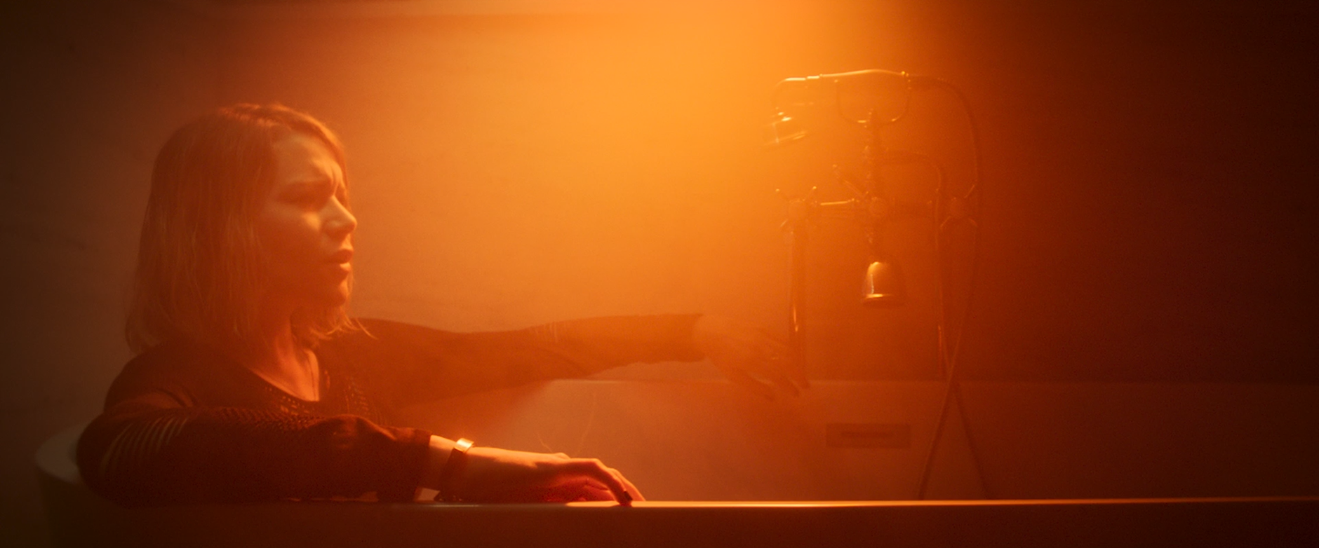 A woman with shoulder-length hair sitting at a piano, reaching out with her right arm, in a dimly lit room with orange lighting and a microphone stand nearby.