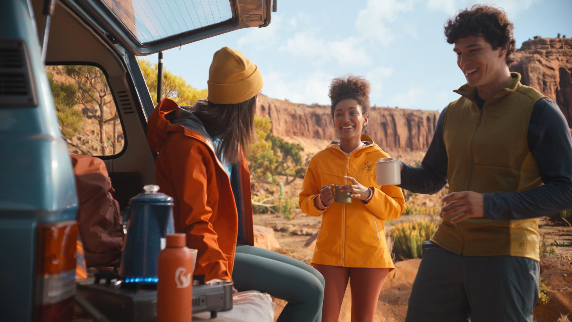 Four people are outdoors near a camping trailer, enjoying hot drinks in a desert landscape with red rock formations and trees in the background.